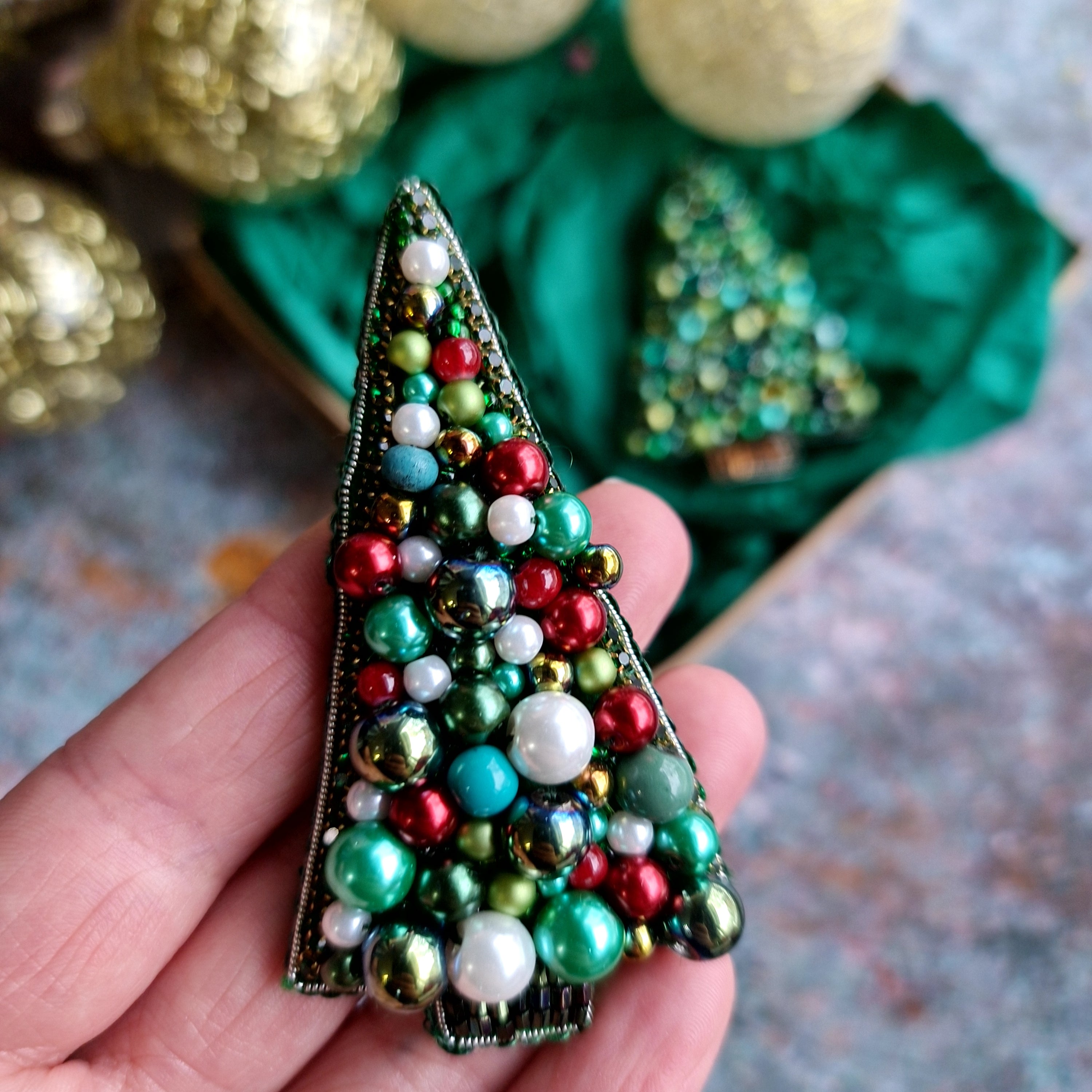 Decorative Christmas tree brooch with colorful beads held in a hand, with more brooches and gold ornaments in the background.