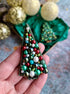 Decorative Christmas tree brooch with colorful beads held in a hand, with more brooches and gold ornaments in the background.