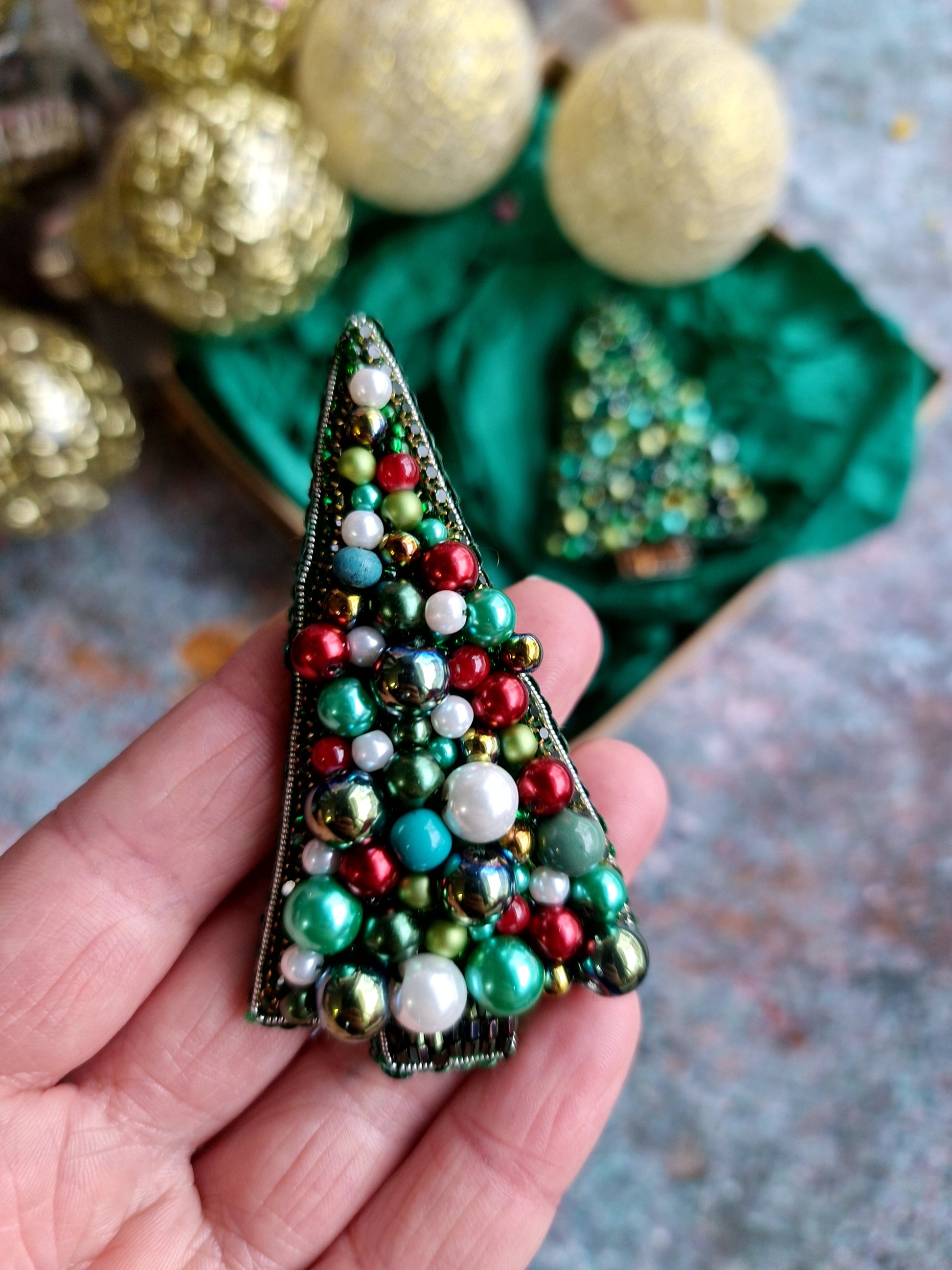 Decorative Christmas tree brooch with colorful beads held in a hand, with more brooches and gold ornaments in the background.