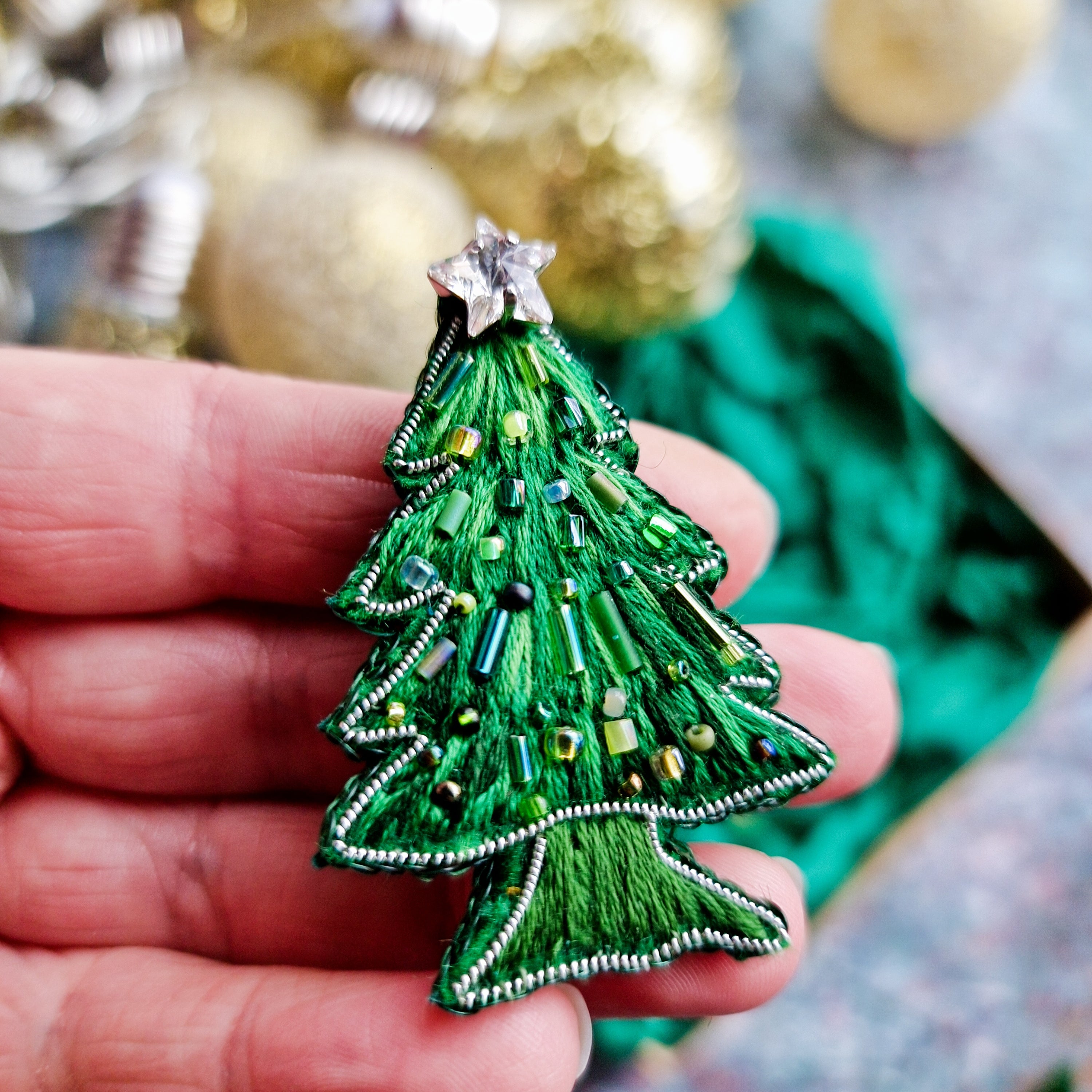 Hand holding a green beaded Christmas tree ornament with gold baubles in the background.