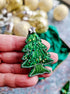 Hand holding a green beaded Christmas tree ornament with gold baubles in the background.