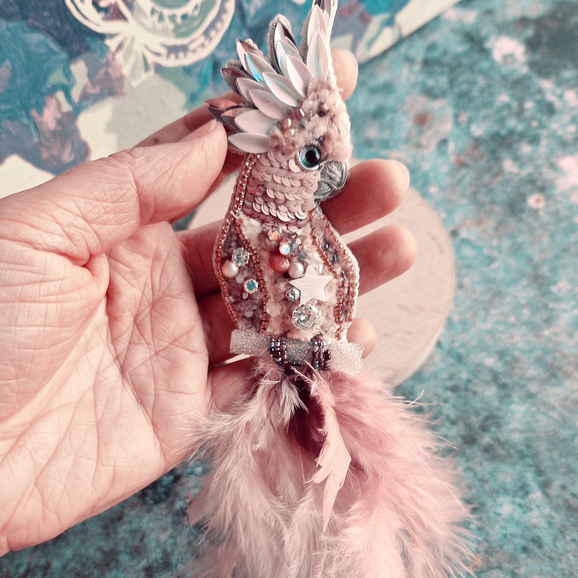 Decorative fish ornament with pink feathers held by a hand on a textured surface.