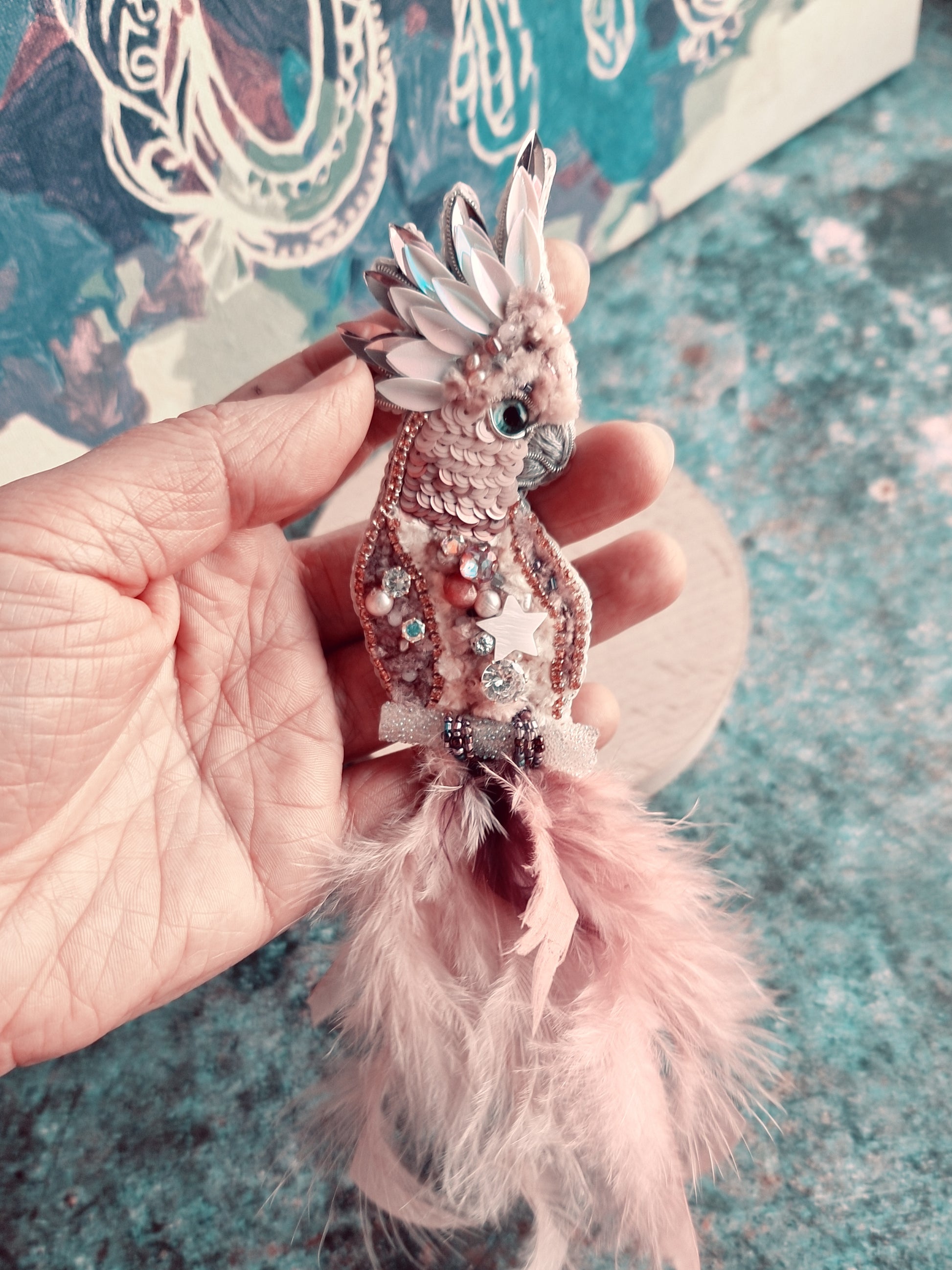 Decorative fish ornament with pink feathers held by a hand on a textured surface.