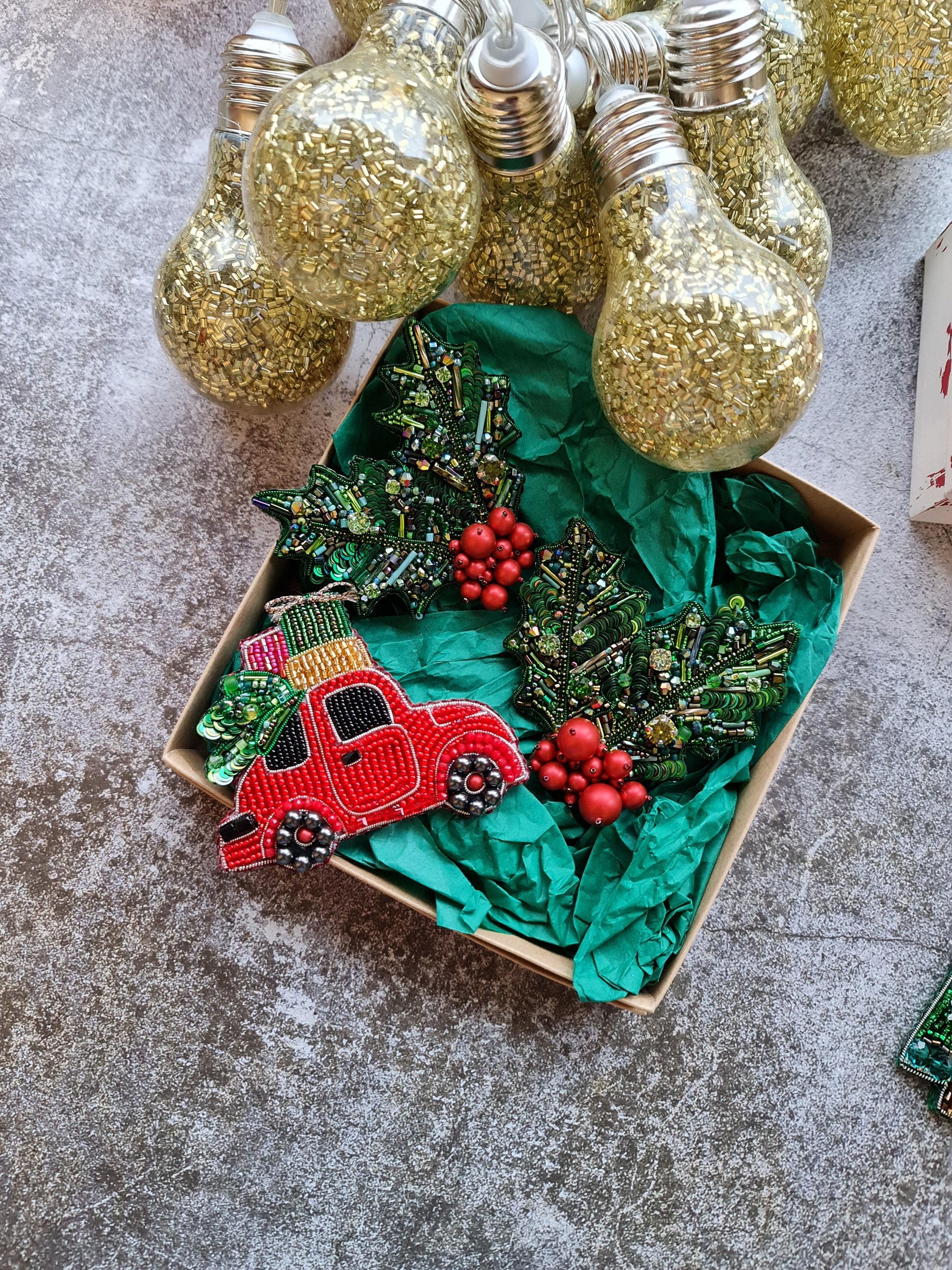 Decorative Christmas wreath with red car and holly berries in a box, surrounded by gold light bulbs on a textured surface.