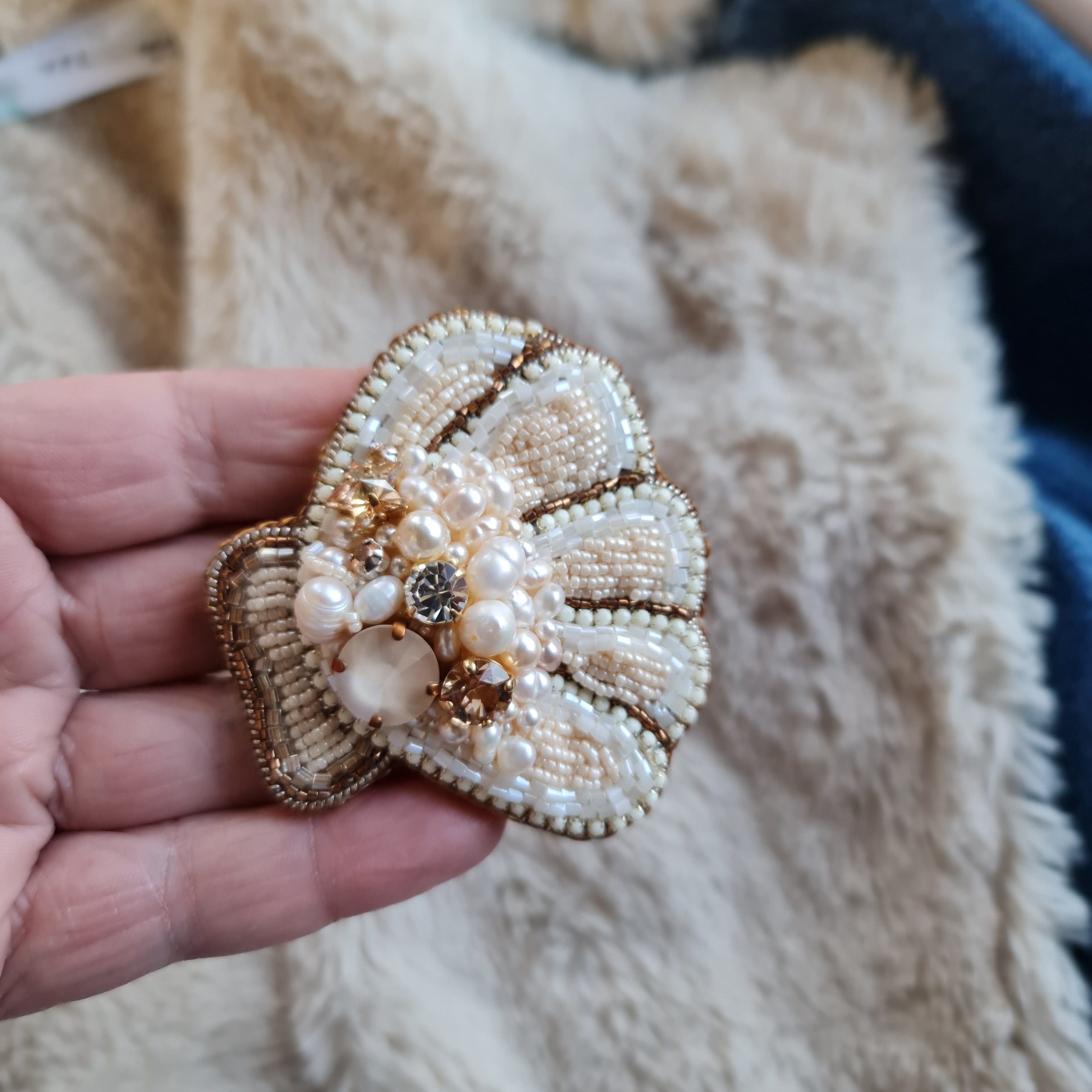 Hand holding a decorative shell brooch with pearls and gold details on a textured surface.
