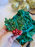 Decorative green holly brooch with red berries held by a hand, with green fabric and gold ornaments in the background.