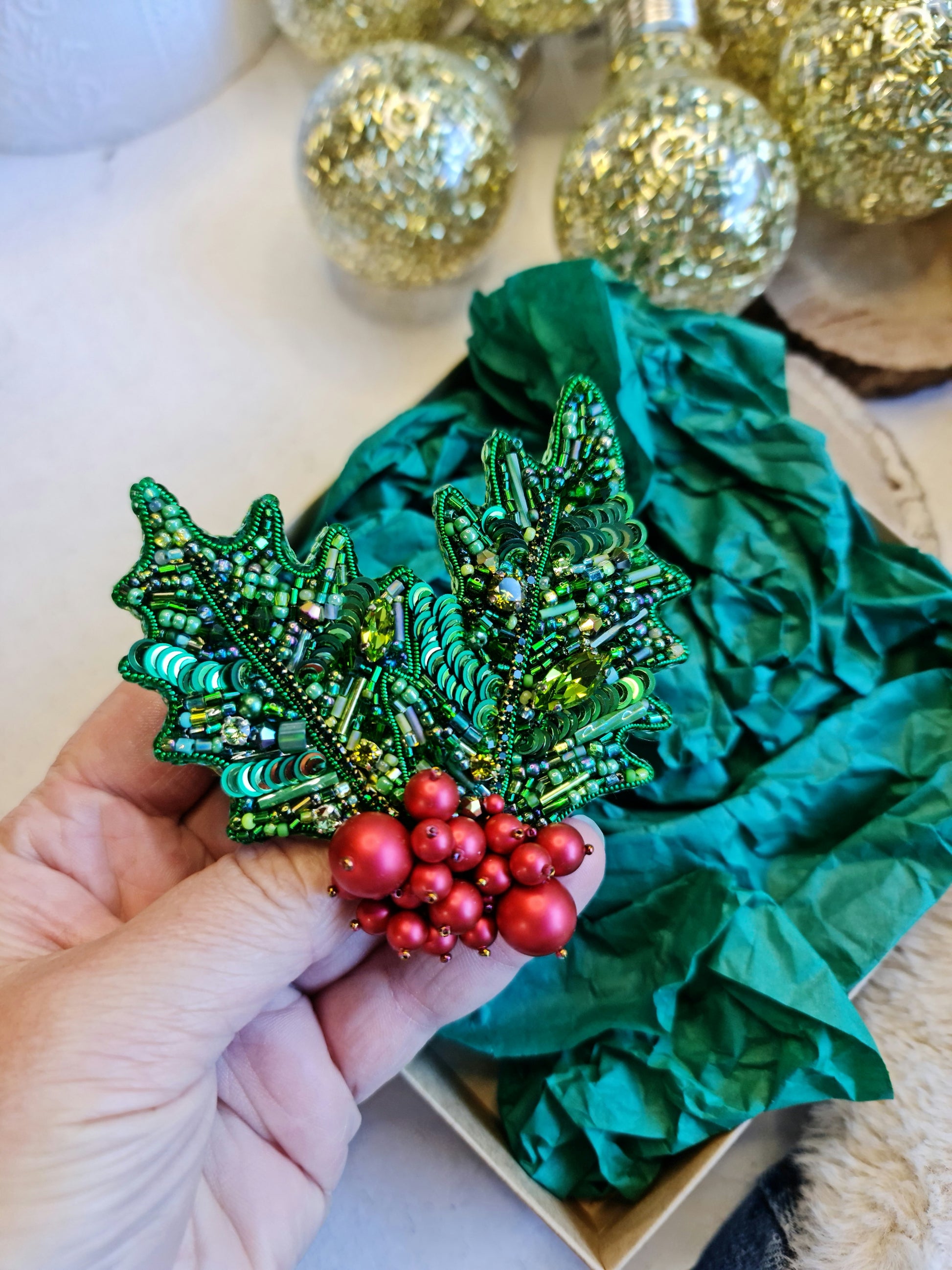 Decorative green holly brooch with red berries held by a hand, with green fabric and gold ornaments in the background.