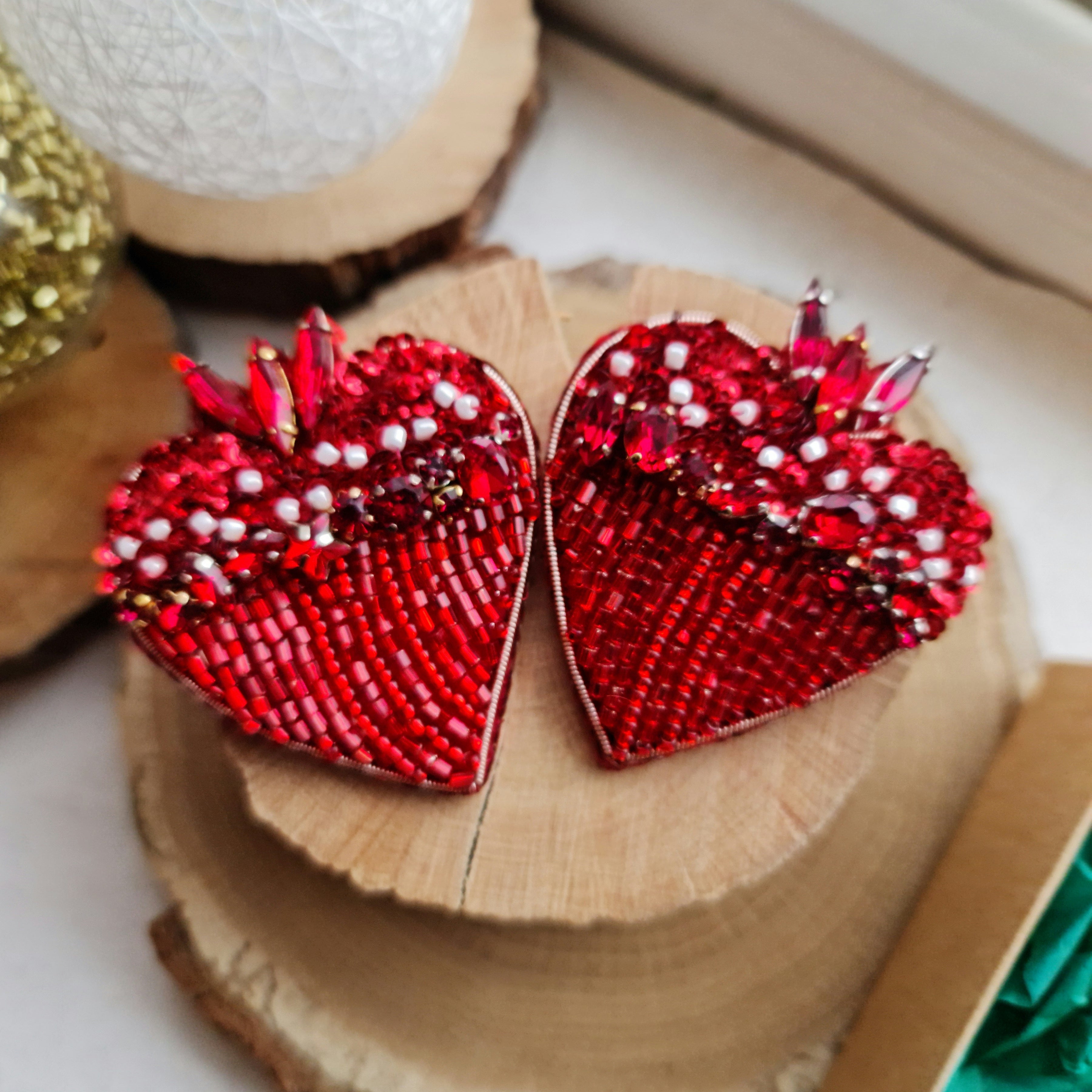 A pair of red heart-shaped beaded earrings with rhinestone accents, displayed on stacked natural wood slices.