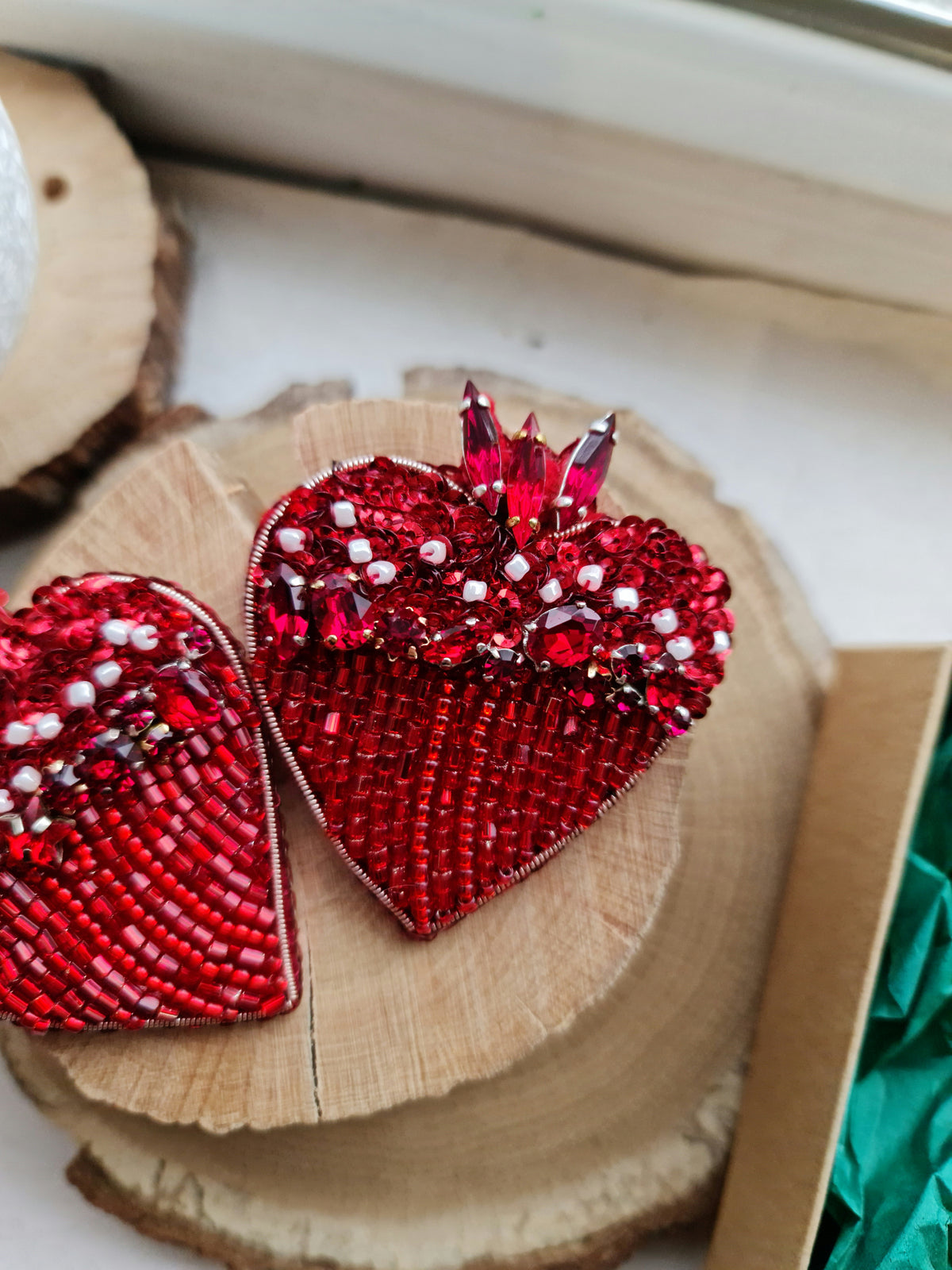 Two red beaded heart-shaped decorative pieces displayed on a round wood slice, with one piece featuring a cluster of faceted red crystals at the top.