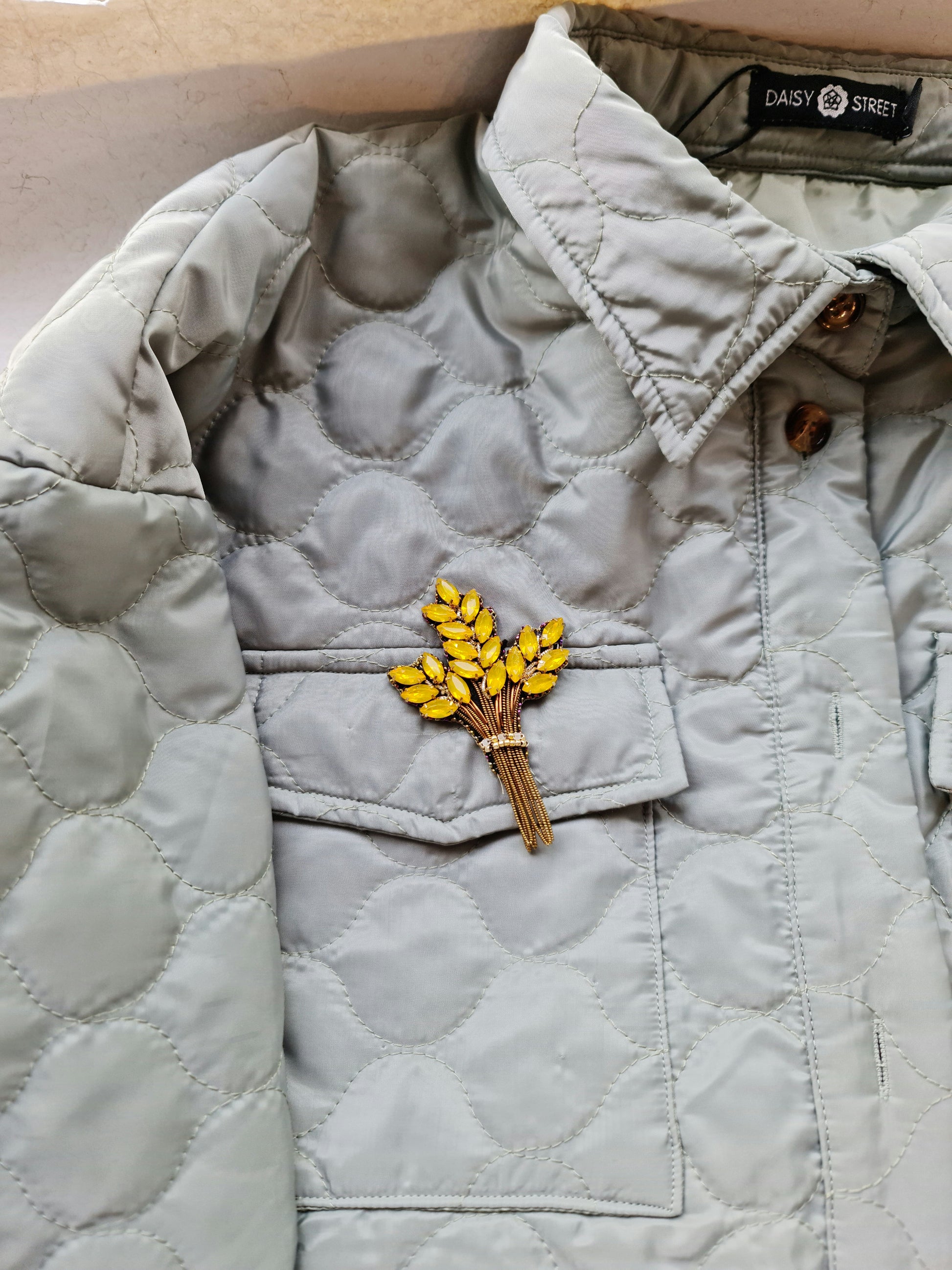 Light gray quilted jacket with a brooch featuring yellow flowers on a matching background