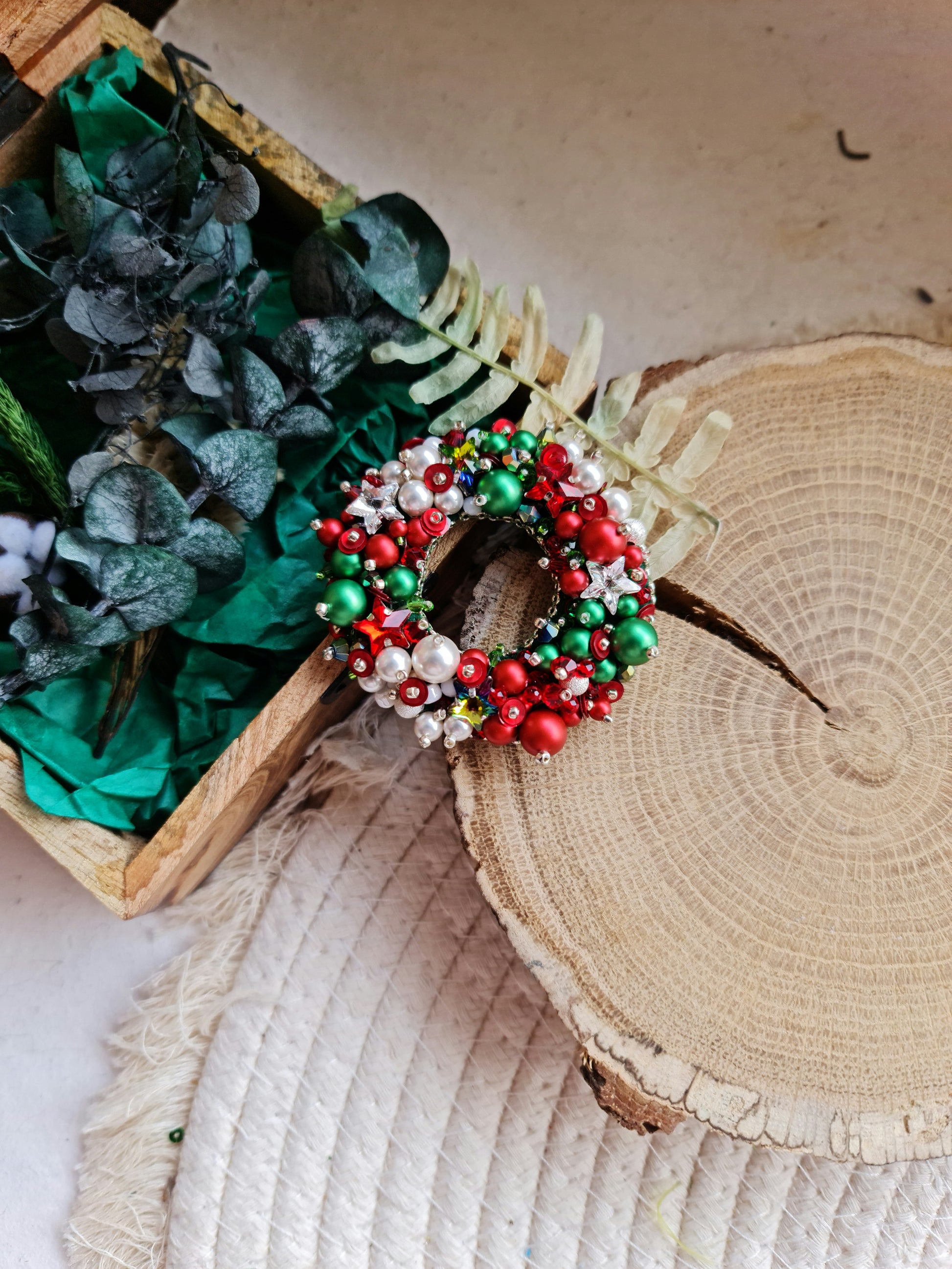 Decorative wreath with red, green, and white beads on a wooden surface with greenery.