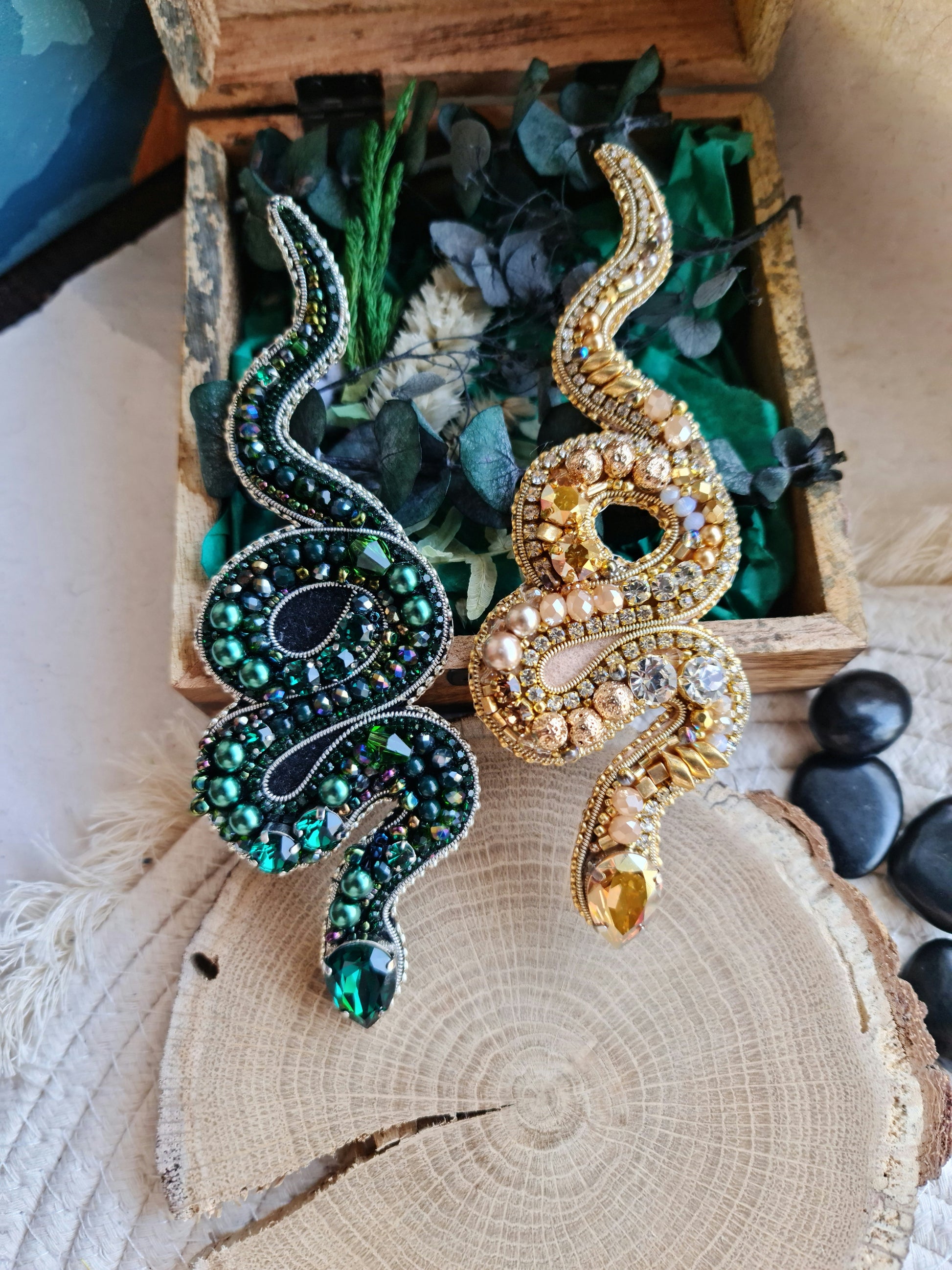 Two decorative snake-shaped pins, one green and one gold, on a wooden surface with greenery.