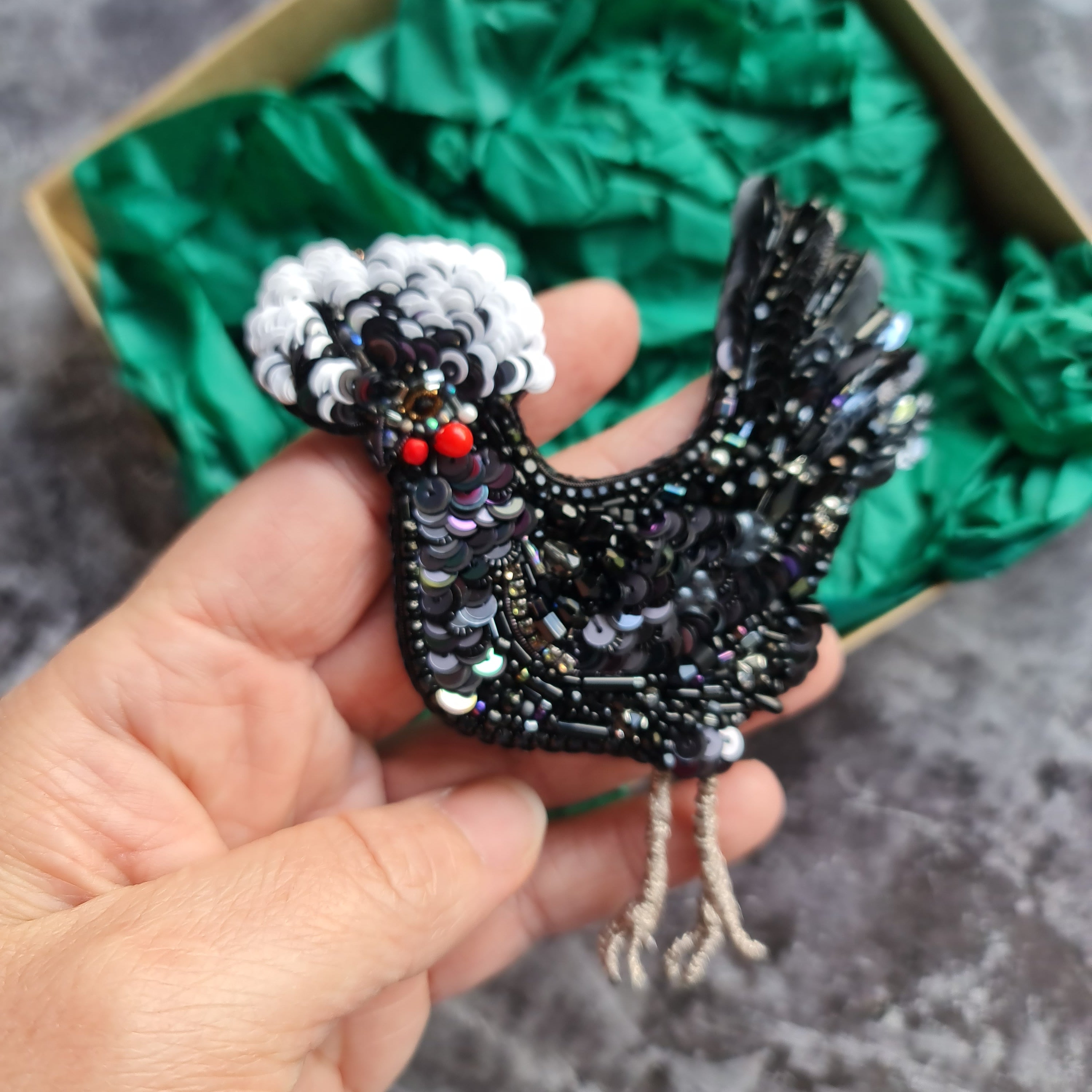 Hand holding a decorative black bird ornament with gemstones against a green textured box.