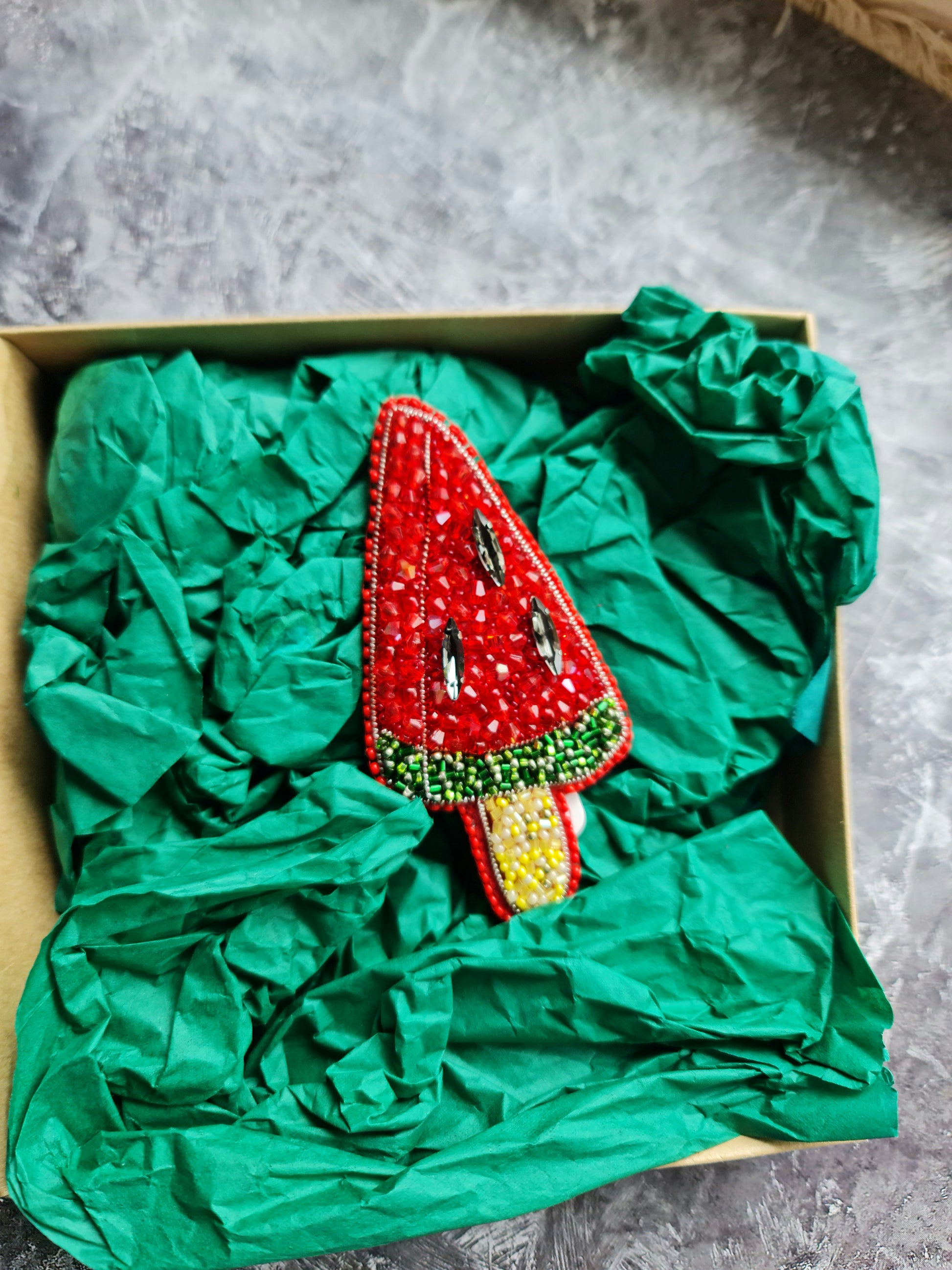 Decorative watermelon-shaped item in a box with green tissue paper on a gray surface