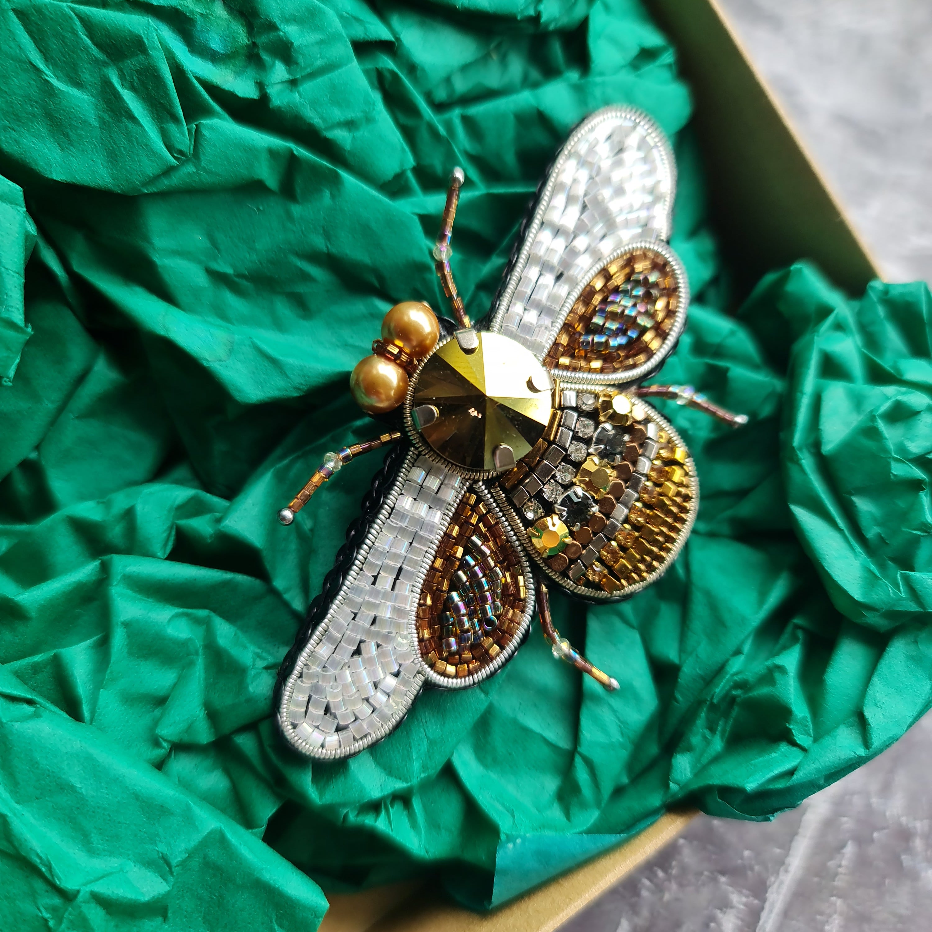 Decorative bee-shaped brooch with gold and silver details on green tissue paper.