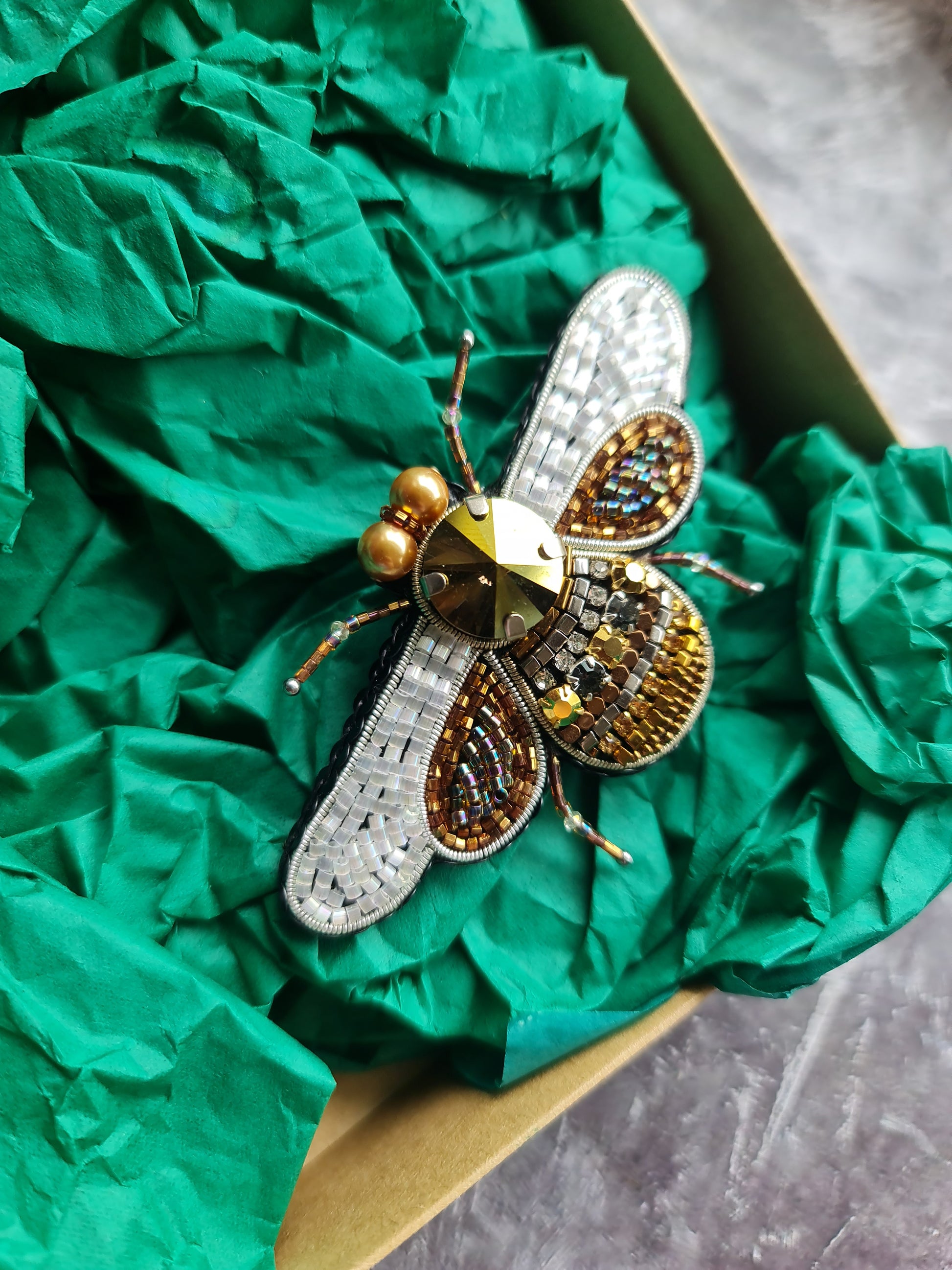 Decorative bee-shaped brooch with gold and silver details on green tissue paper.
