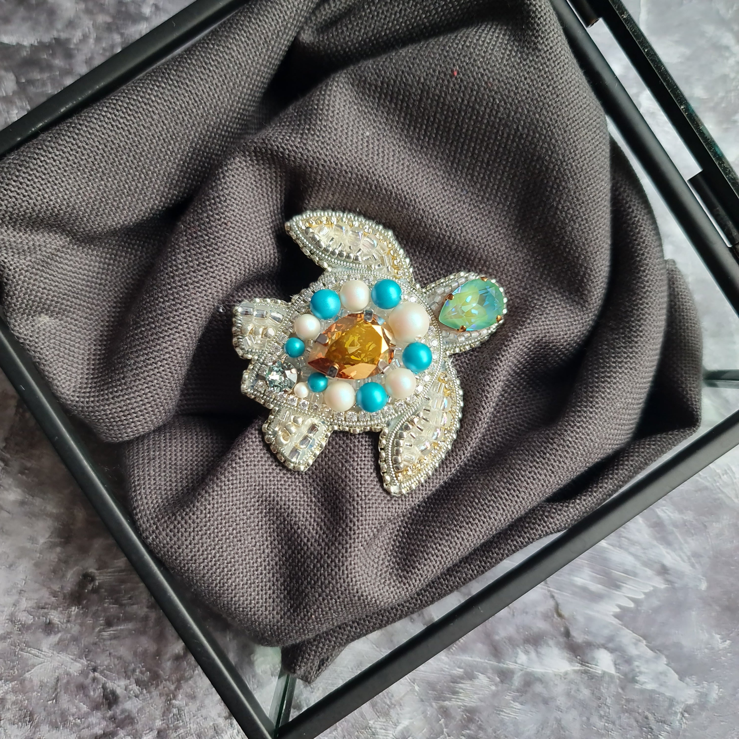 Decorative turtle-shaped object with gemstones on a gray fabric inside a glass box.