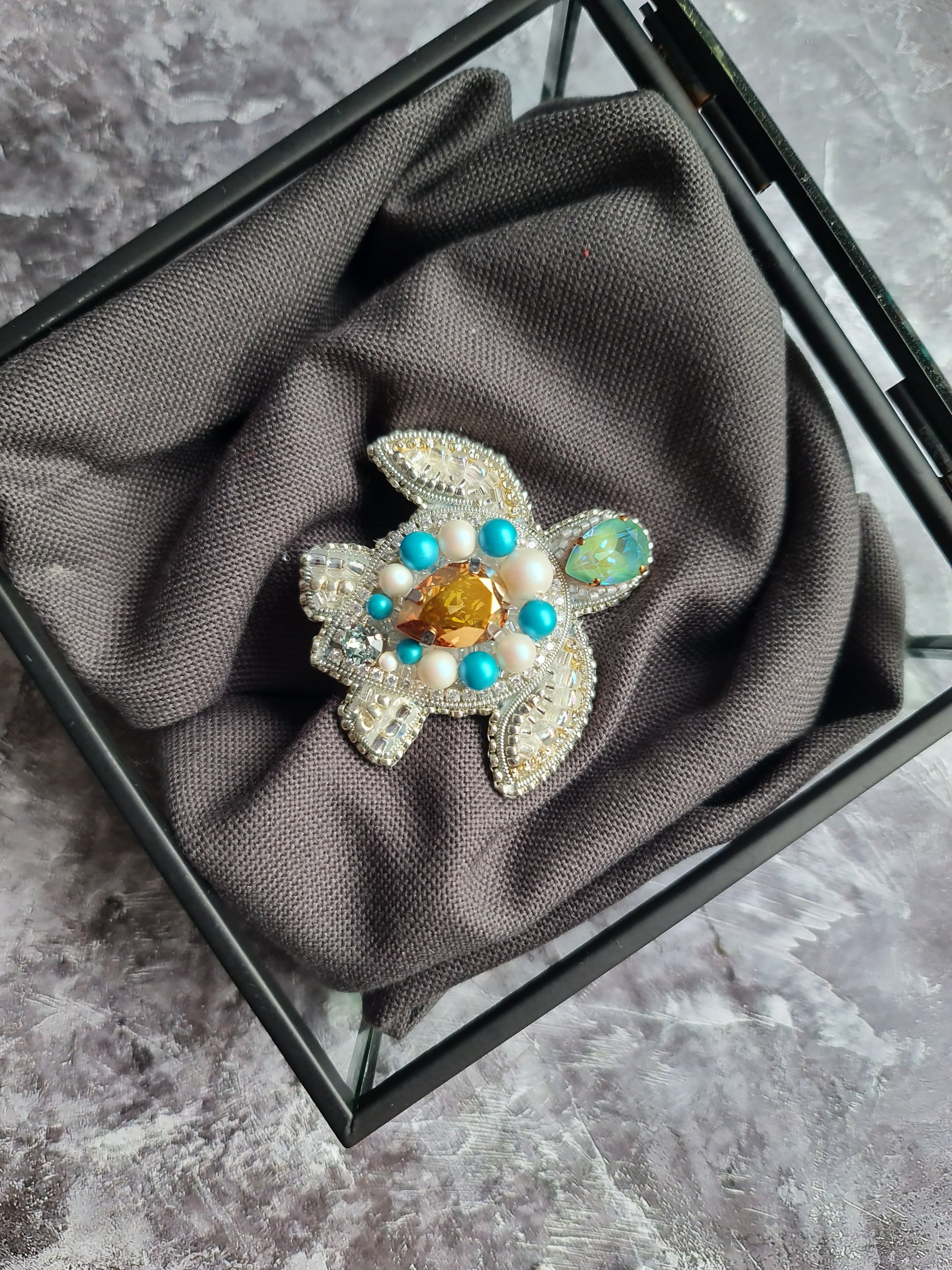Decorative turtle-shaped object with gemstones on a gray fabric inside a glass box.
