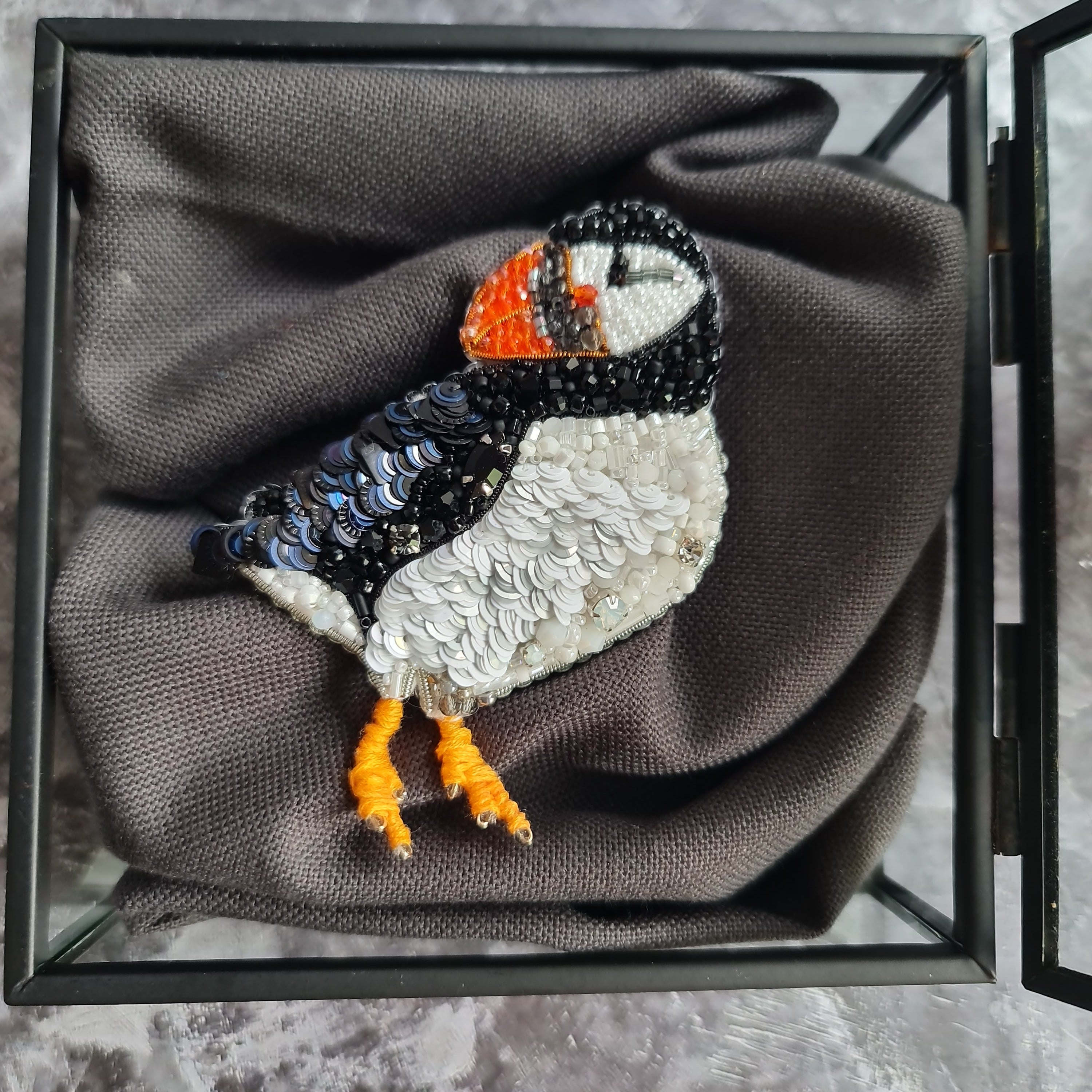Penguin-shaped brooch in a glass display box on a textured gray surface