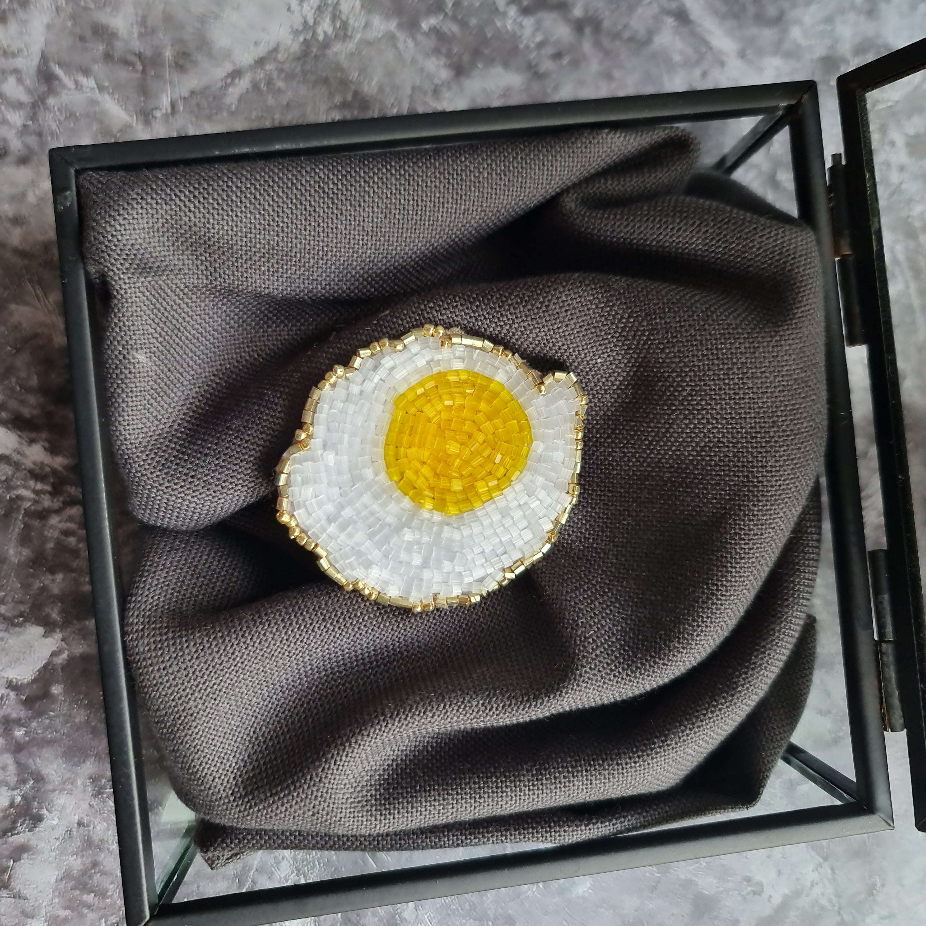 Brooch shaped like a fried egg in a jewelry box on a marble surface