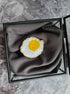 Brooch shaped like a fried egg in a jewelry box on a marble surface