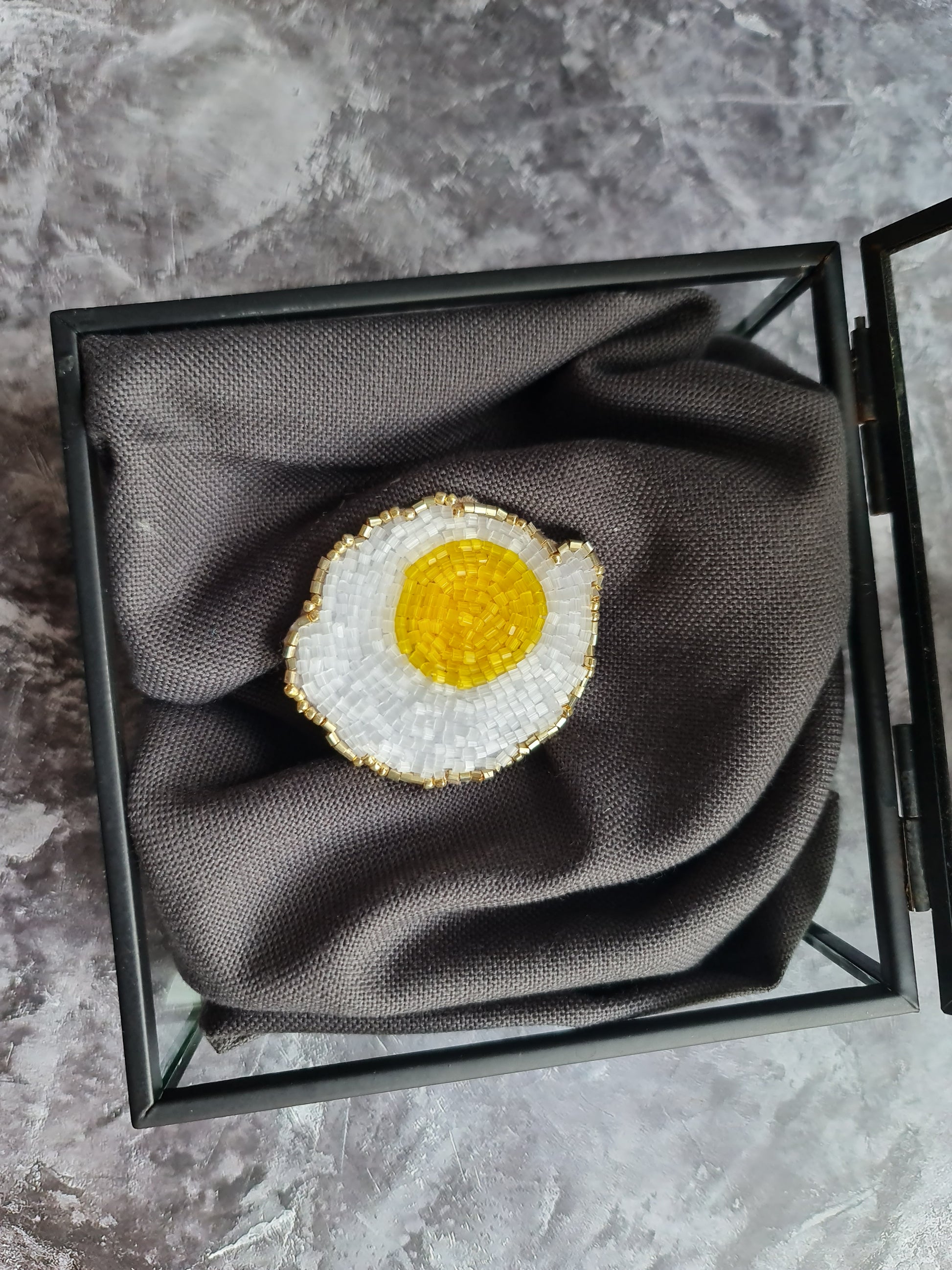 Brooch shaped like a fried egg in a jewelry box on a marble surface