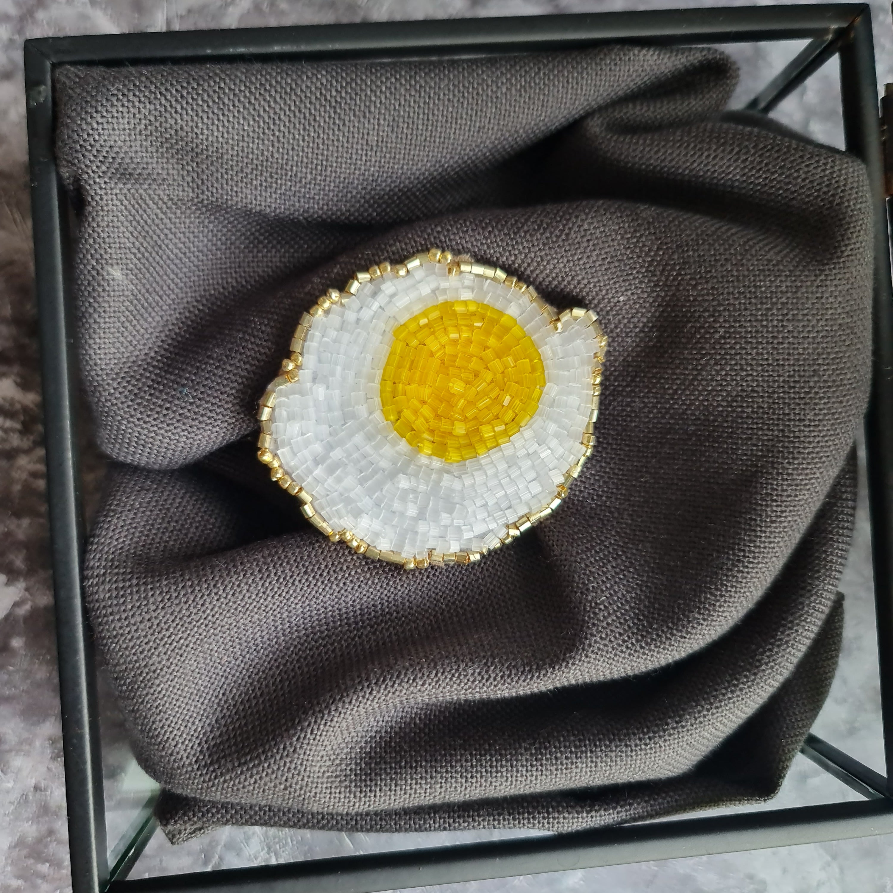 Decorative brooch shaped like a fried egg on a gray fabric in a black frame.