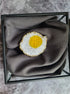 Decorative brooch shaped like a fried egg on a gray fabric in a black frame.