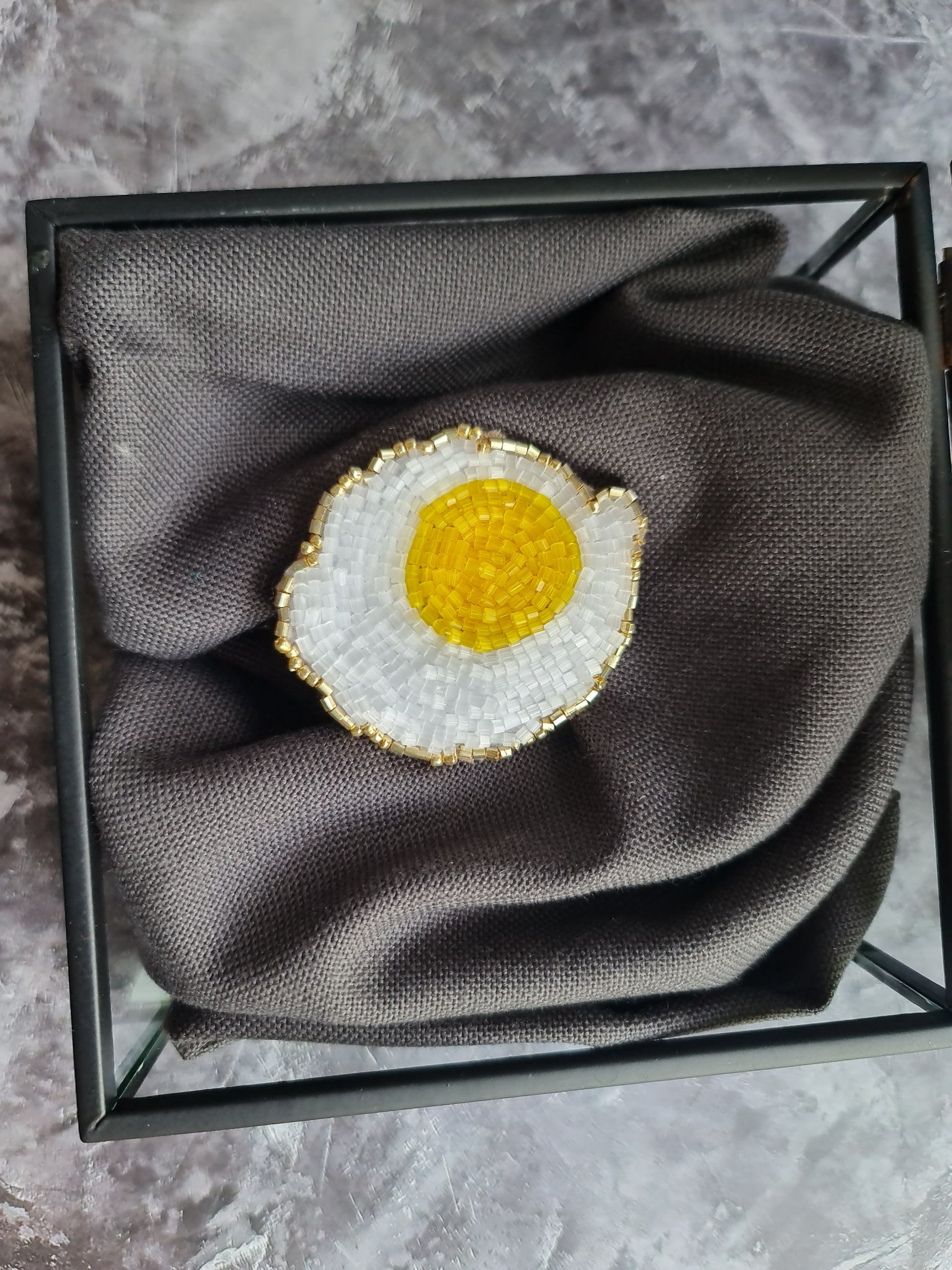 Decorative brooch shaped like a fried egg on a gray fabric in a black frame.
