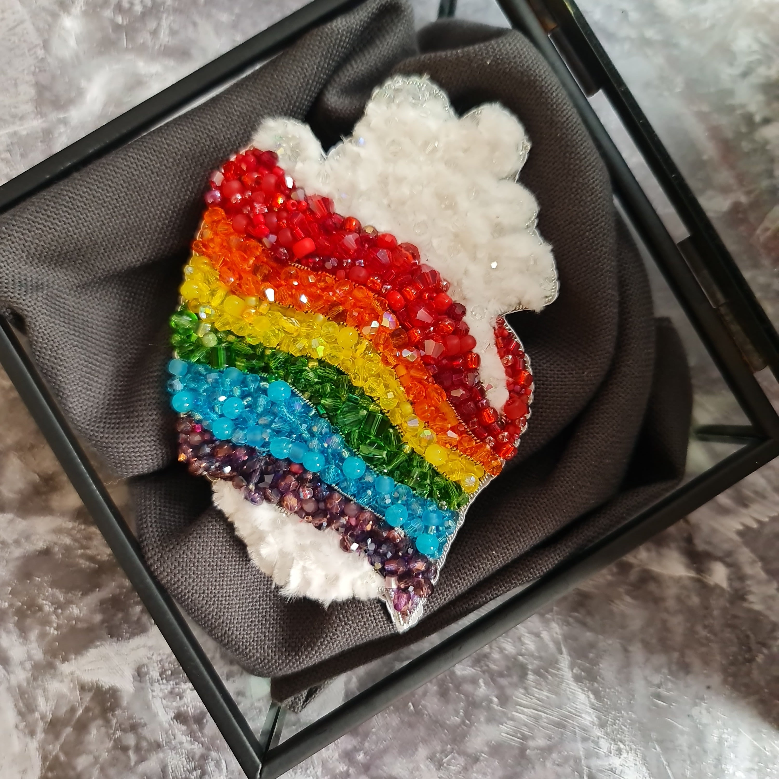 Rainbow-colored brooch with crystals in a black display case on a textured surface