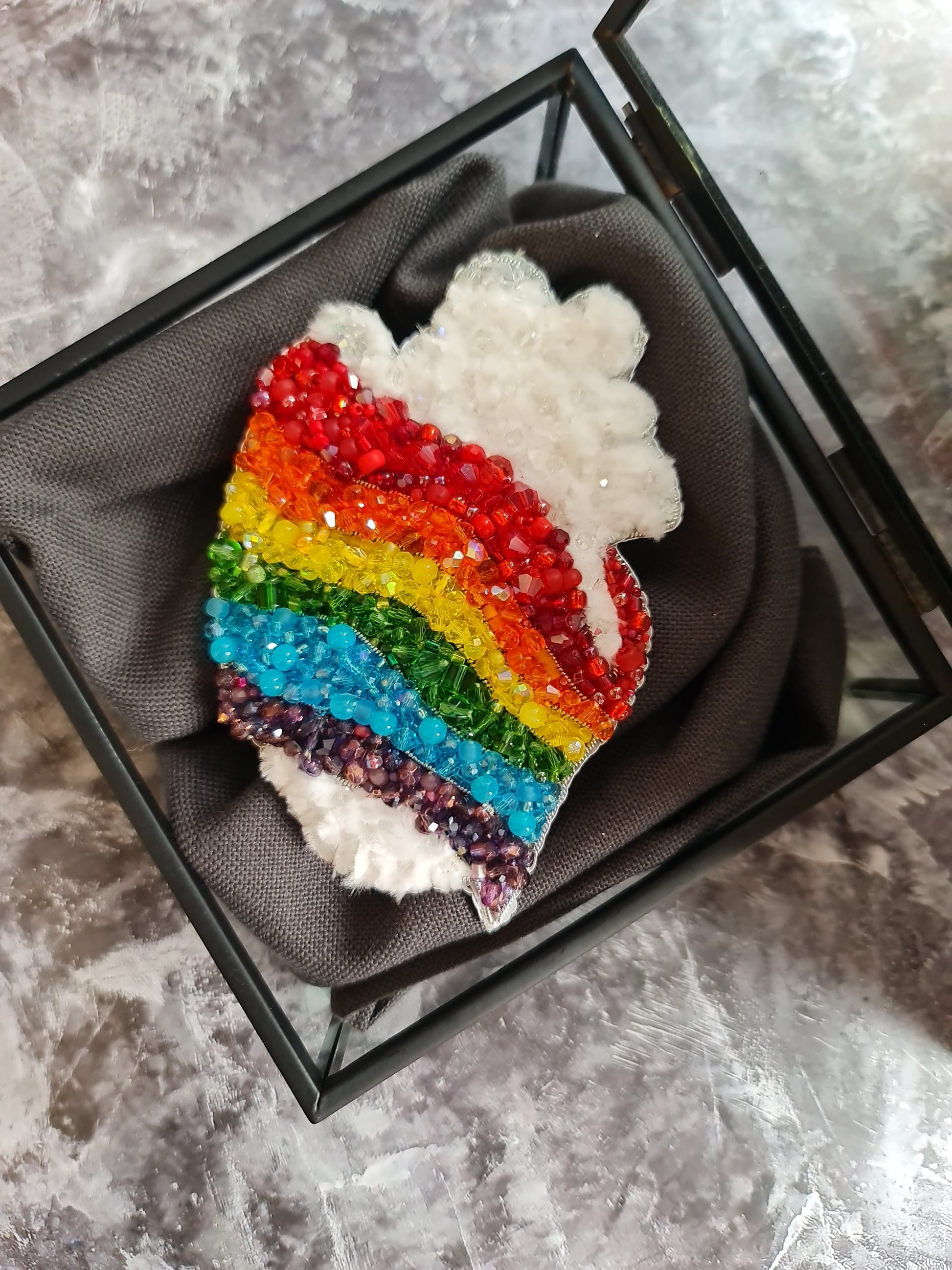 Rainbow-colored brooch with crystals in a black display case on a textured surface
