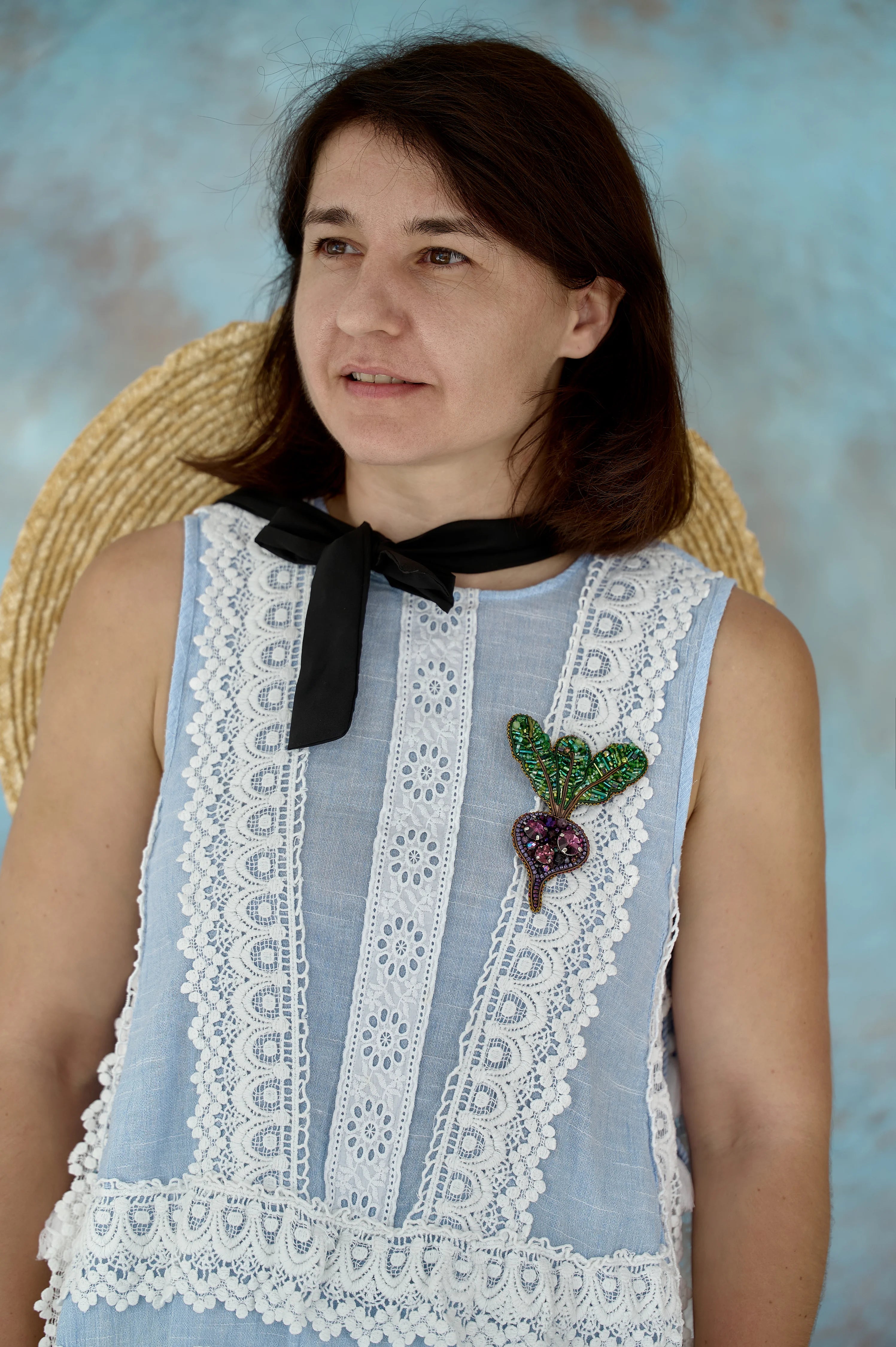 Woman wearing a light blue sleeveless top with lace details and a brooch, sitting against a textured wall.