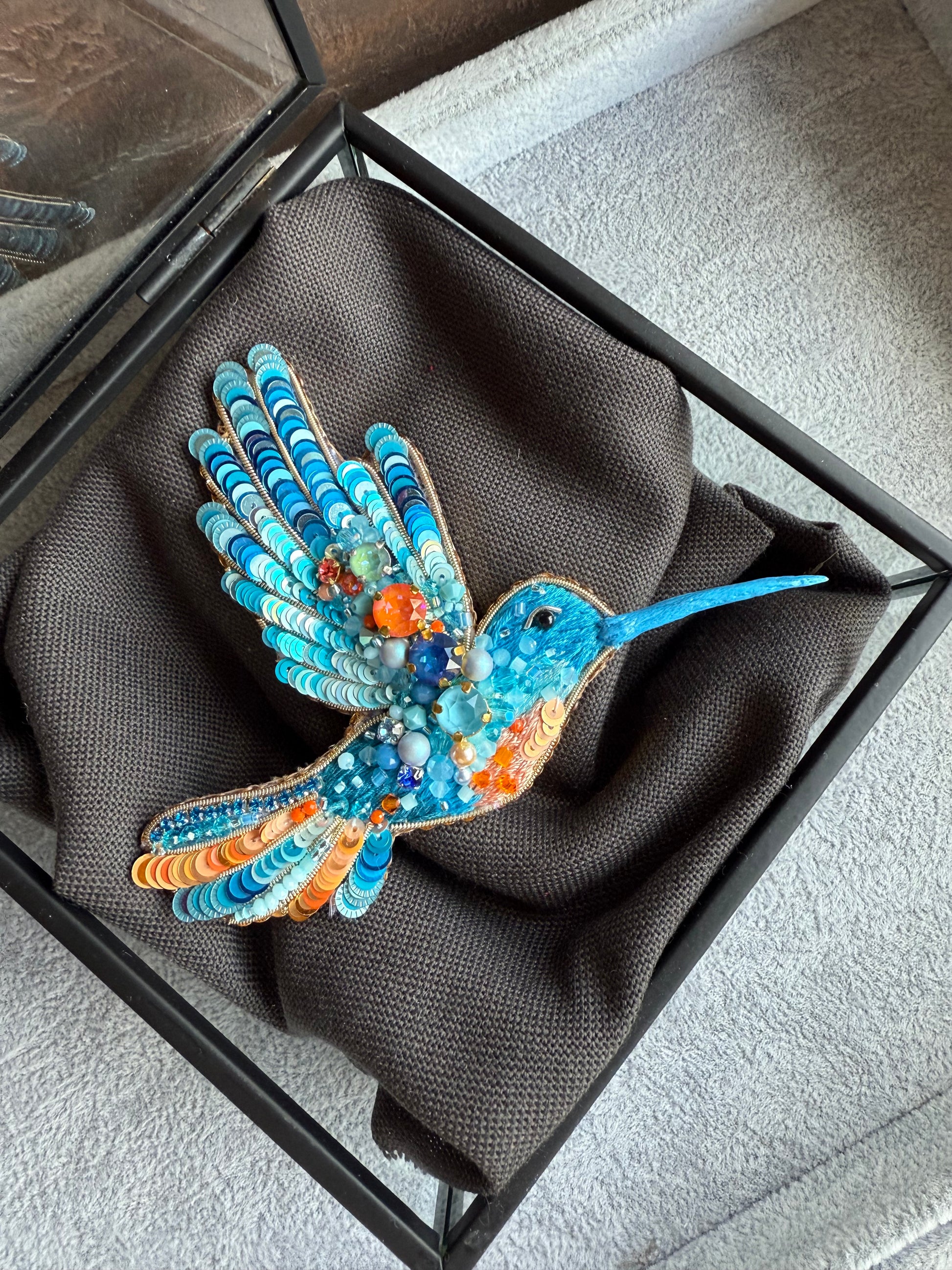 Colorful hummingbird brooch in a black box on a gray surface