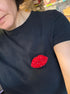 A close-up of a person wearing a black short-sleeve t-shirt with a red glittery lip-shaped patch on the chest, set against a cluttered background of storage bins.