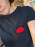 A close-up selfie of a person wearing a black t-shirt with a red glittery lip-shaped embellishment on the chest, set against a cluttered indoor background.
