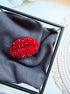 A red lip-shaped brooch embellished with rhinestones, displayed on a dark gray textured fabric inside a black square frame.