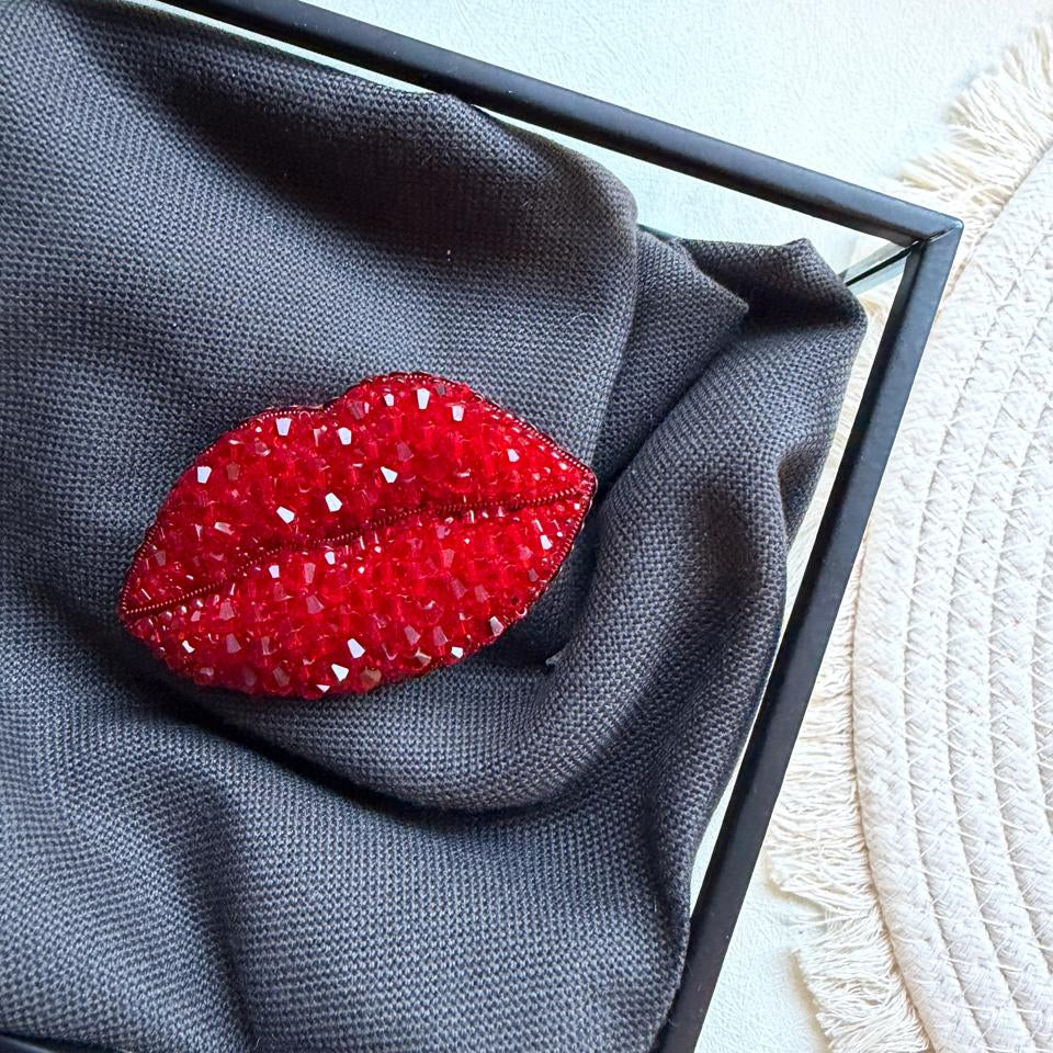 A red lip-shaped brooch embellished with rhinestones, displayed on a dark gray textured fabric inside a black square frame.