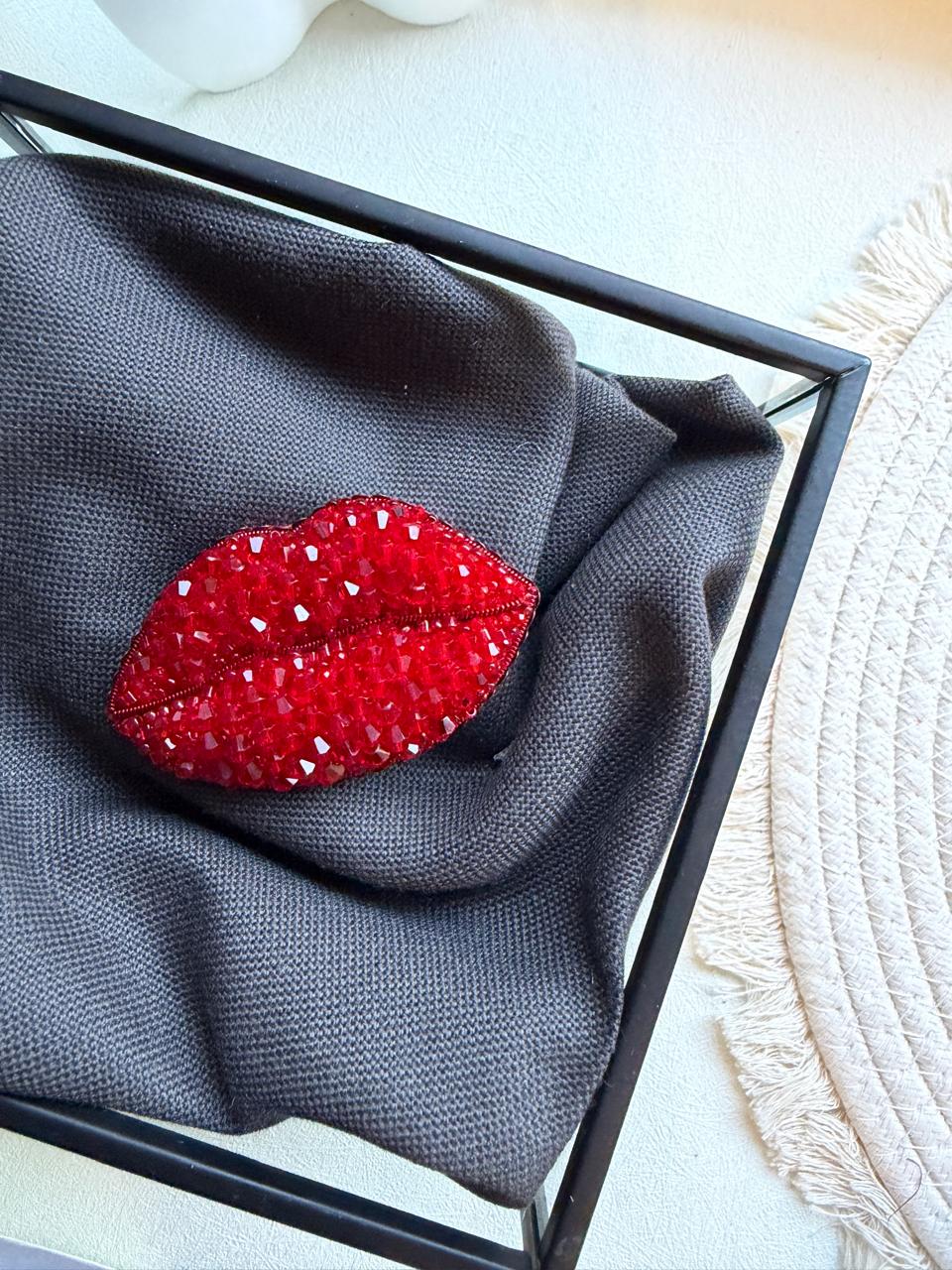A red lip-shaped brooch embellished with rhinestones, displayed on a dark gray textured fabric inside a black square frame.