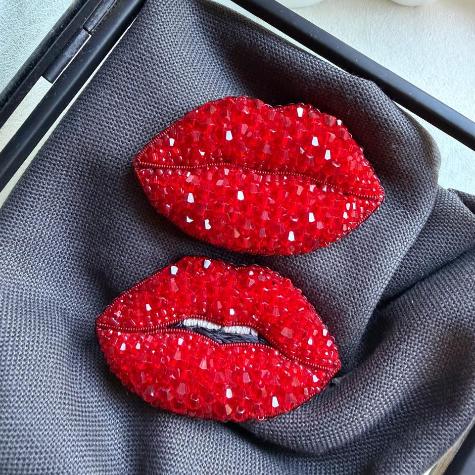 A pair of red lip-shaped brooch pins covered in sparkling rhinestones, displayed on a textured gray fabric insert inside a black jewelry box.