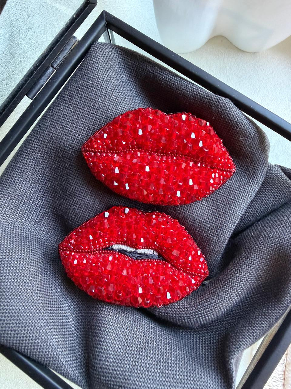 A pair of red lip-shaped brooch pins covered in sparkling rhinestones, displayed on a textured gray fabric insert inside a black jewelry box.
