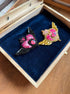 Two decorative winged brooch pins displayed inside an open wooden jewelry box with a dark blue velvet lining. One brooch is black with pink and red details, and the other is gold with pink and red details, both featuring a central pink gem-like motif.
