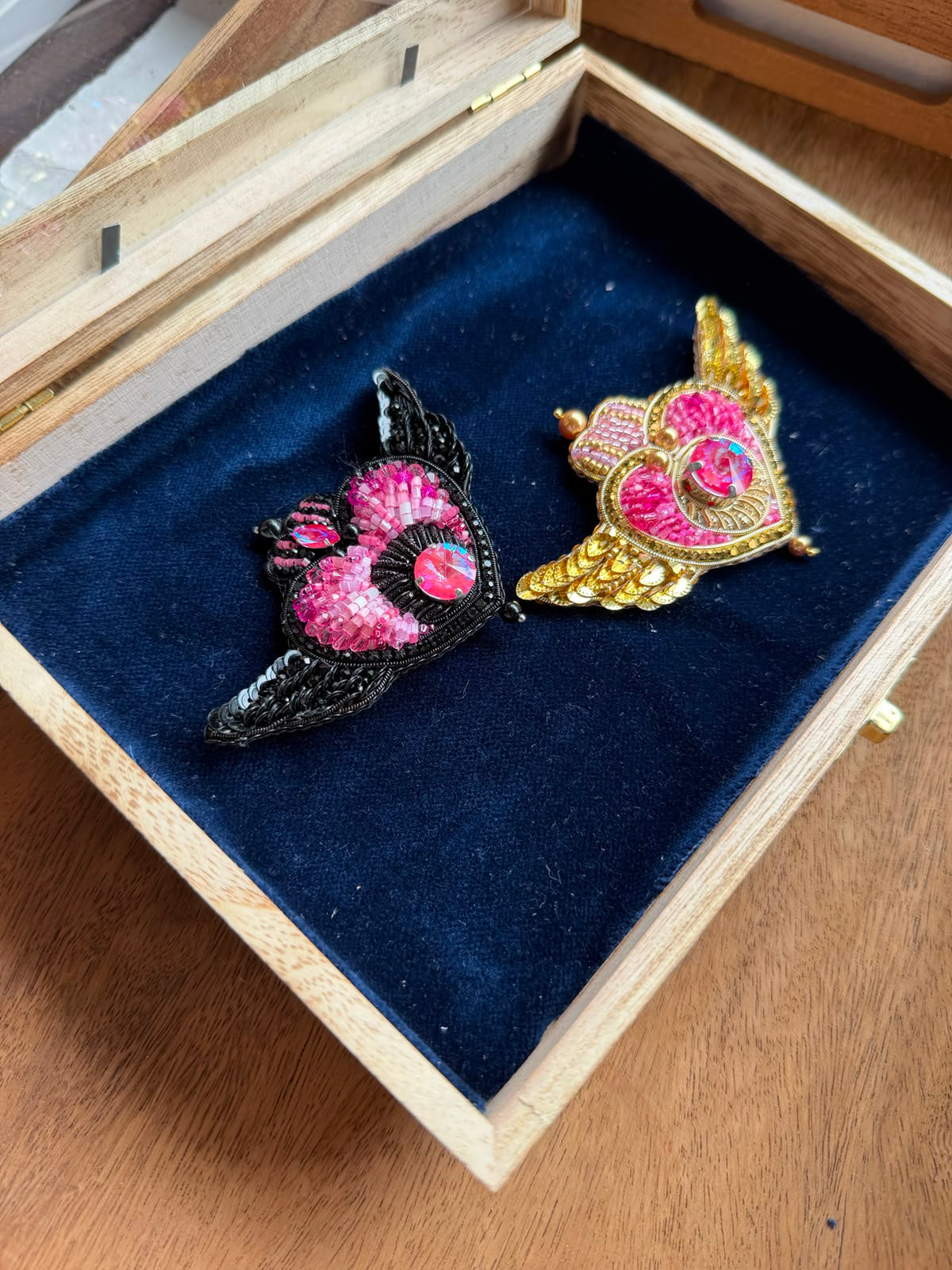 Two decorative winged brooch pins displayed inside an open wooden jewelry box with a dark blue velvet lining. One brooch is black with pink and red details, and the other is gold with pink and red details, both featuring a central pink gem-like motif.