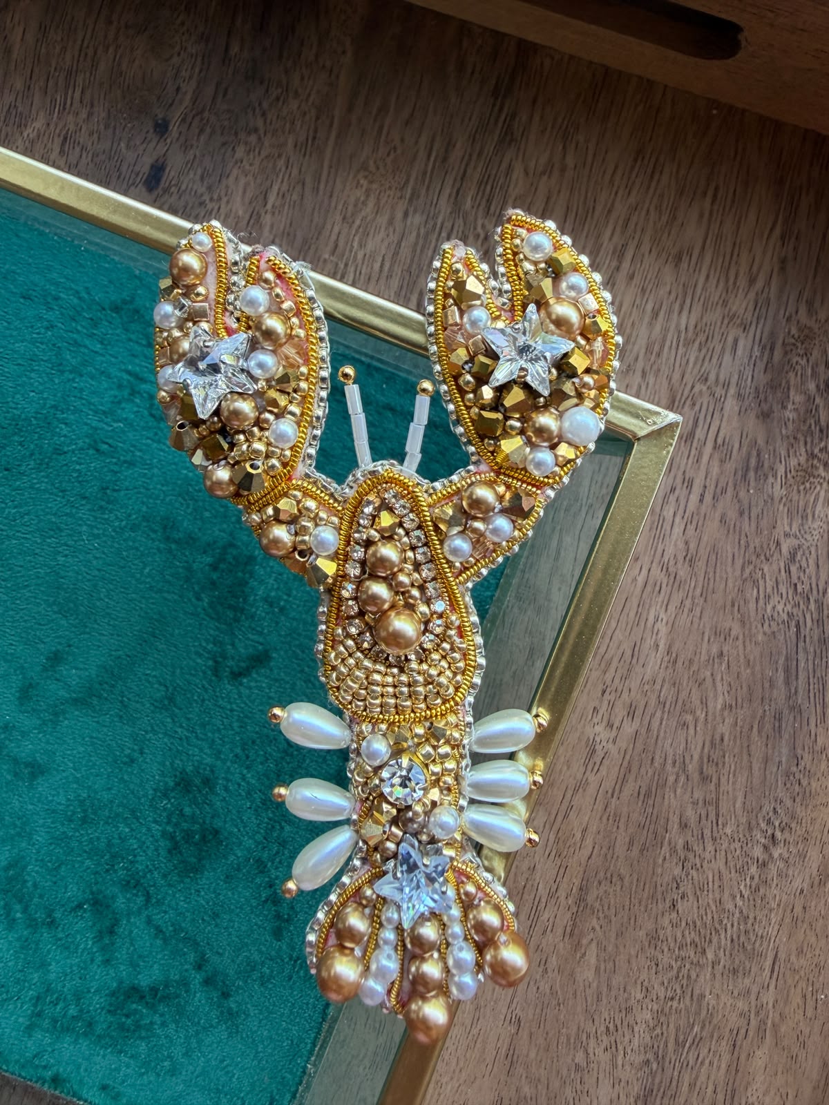 Handmade gold lobster beaded brooch with sparkling star crystals and pearl details, held in hand above a green velvet jewelry box.