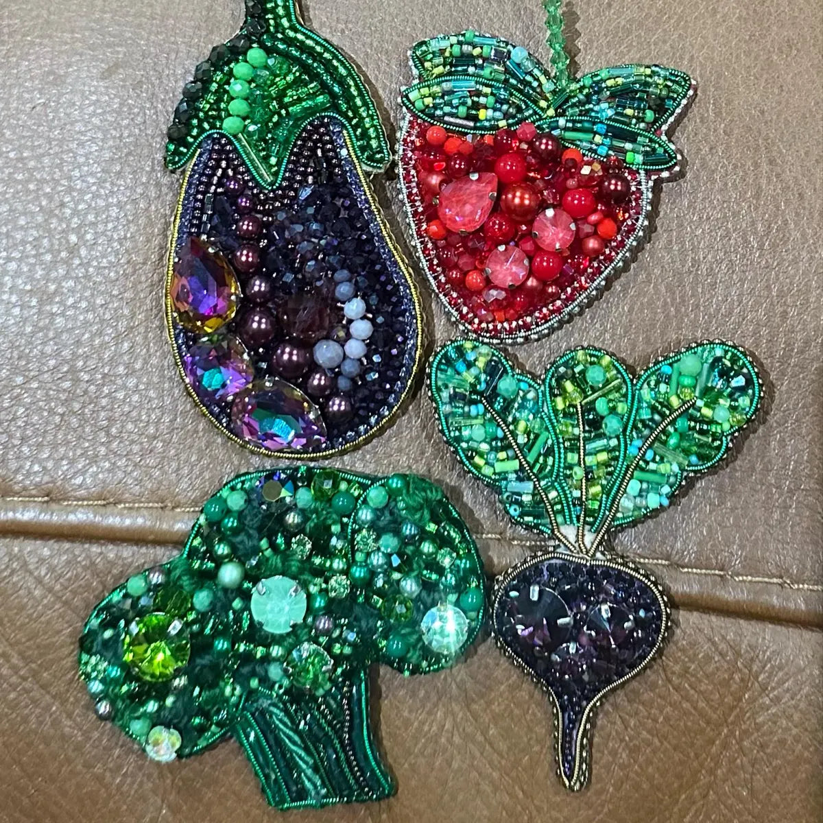 Decorative brooches shaped like an eggplant, strawberry, broccoli, and leaf on a beige background.